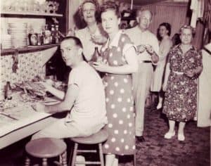 A group of people socializing in a vintage kitchen setting, with one person seated at the table and others standing nearby.
