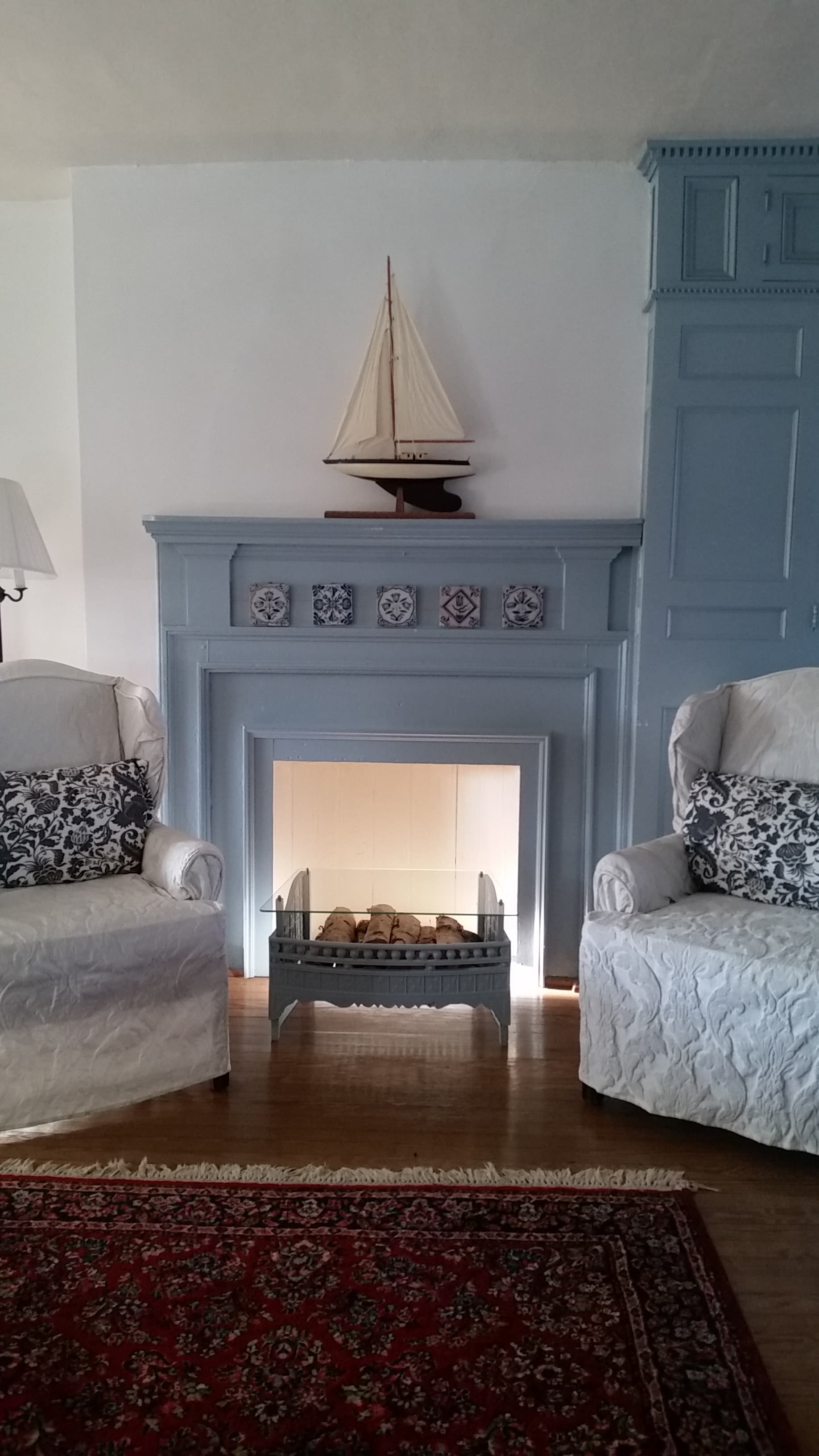A cozy room featuring a blue-painted fireplace, decorative sailboat, two upholstered chairs, and a patterned rug.