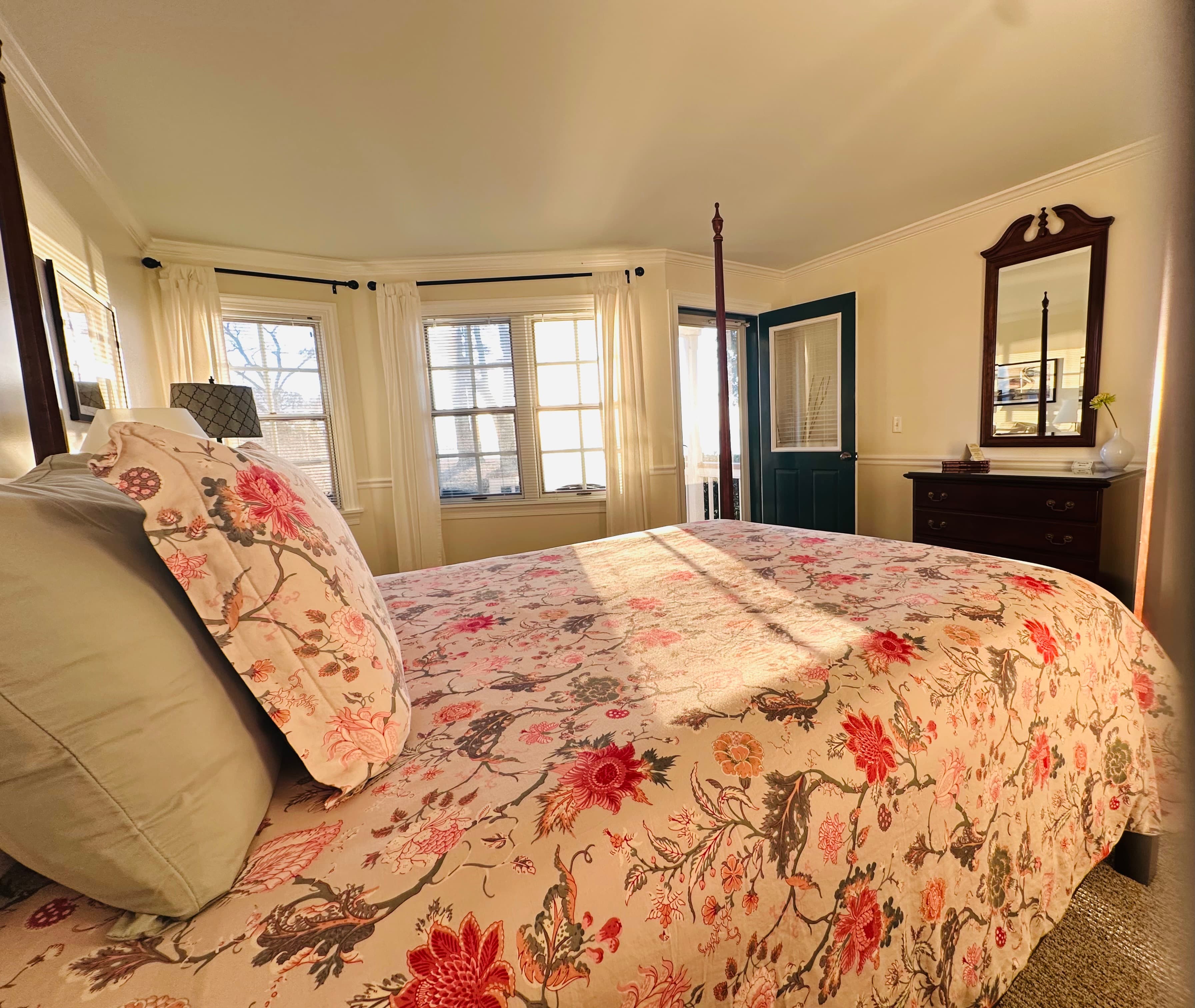 A cozy bedroom with a floral quilt, a mirror, and large windows letting in natural light.