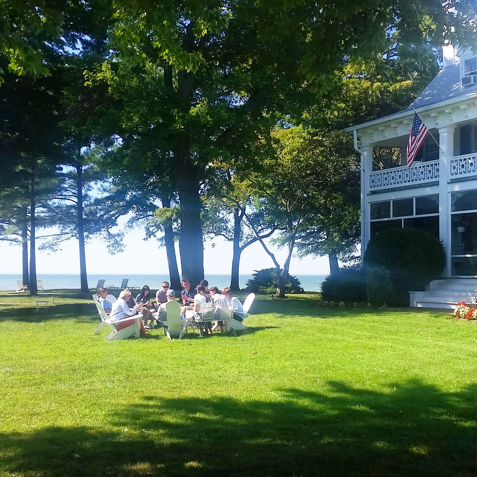 A group of people gathers around a table on a grassy lawn near a lakefront house.