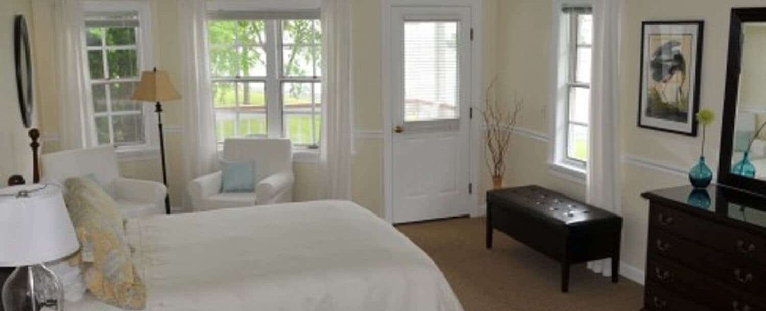 Bright, cozy bedroom featuring a bed, armchair, and dresser, with large windows bringing in natural light.