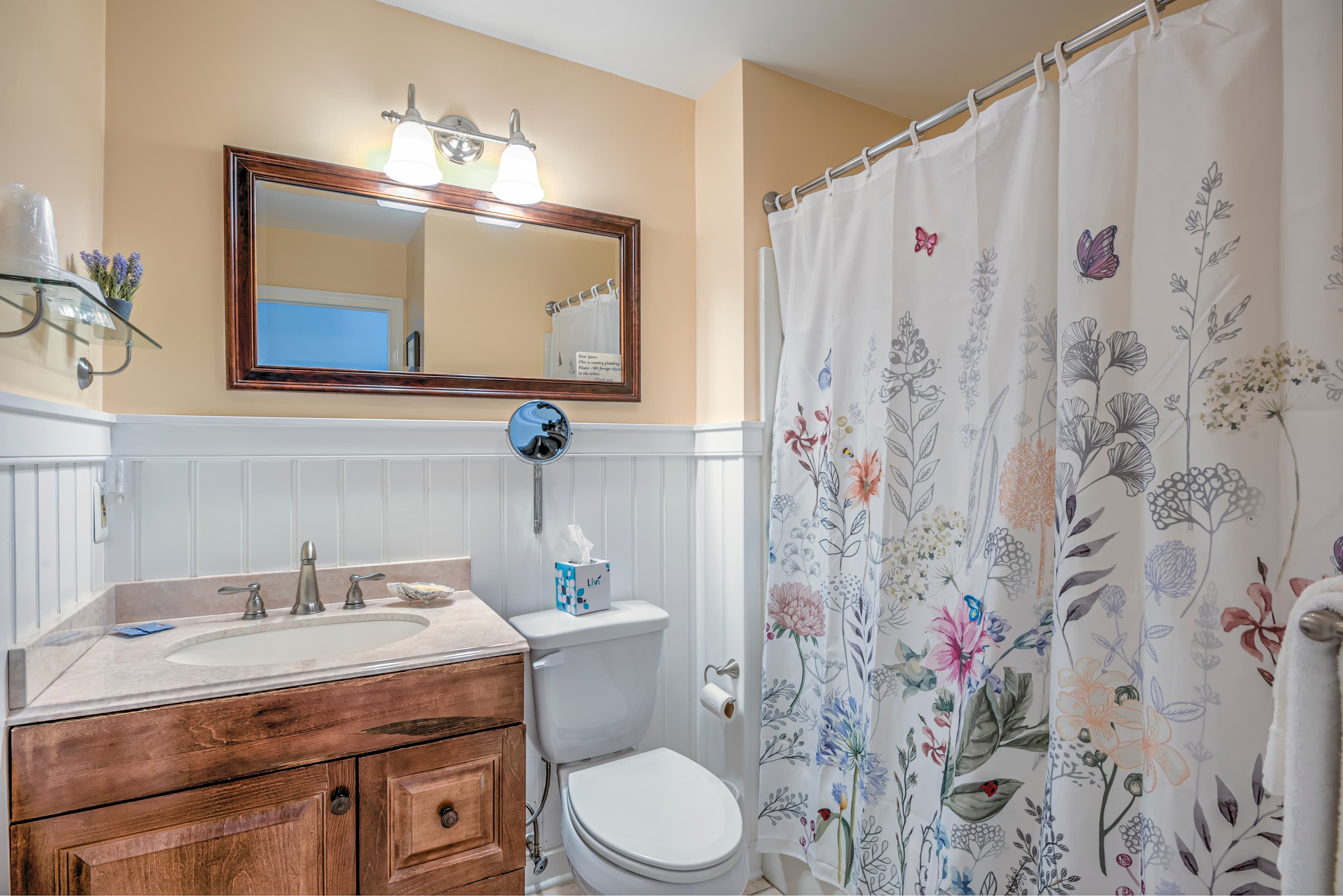 A well-lit bathroom featuring a flower-patterned shower curtain, wooden vanity, and mirror.