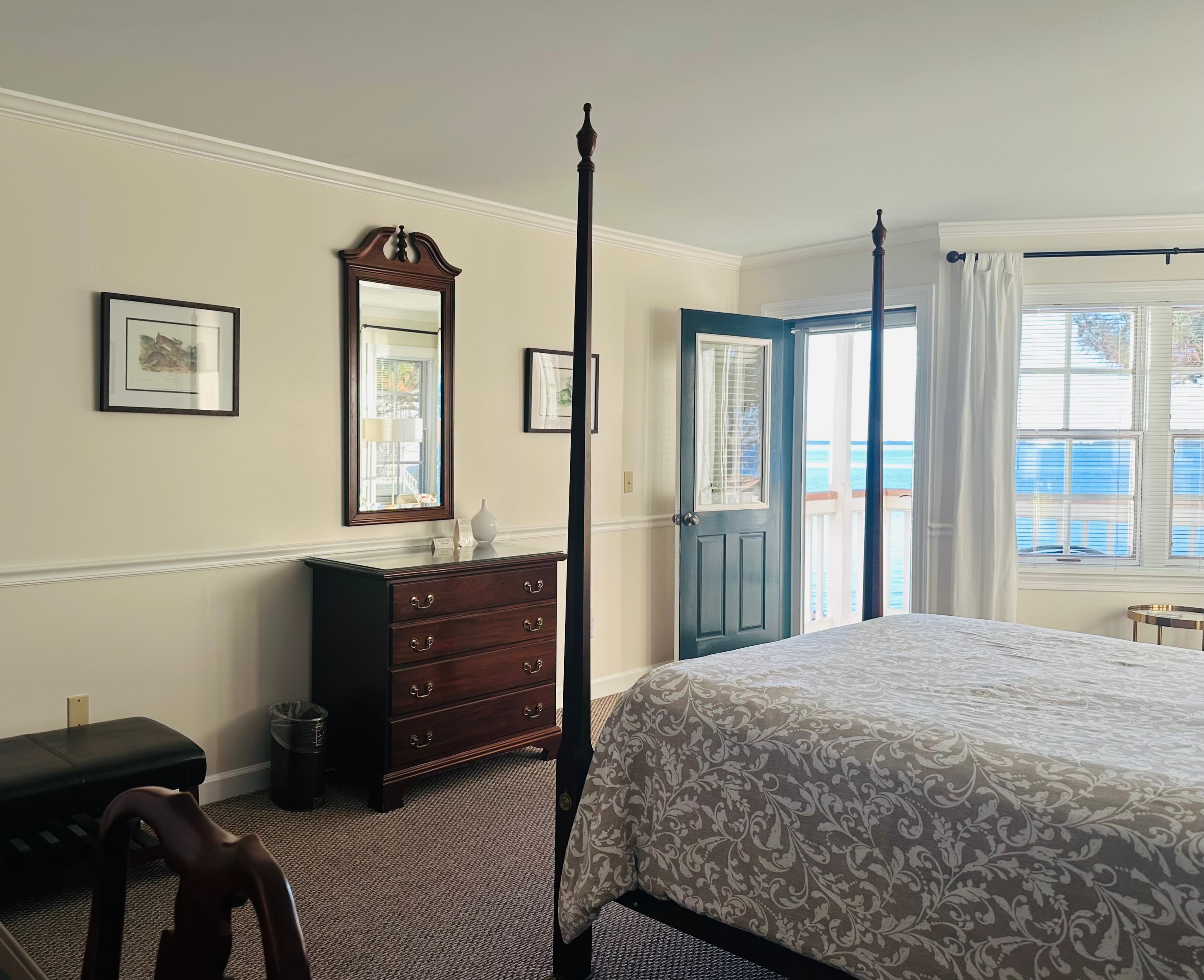 A bright bedroom featuring a four-poster bed, a wooden dresser, a mirror, and a door leading to a balcony.