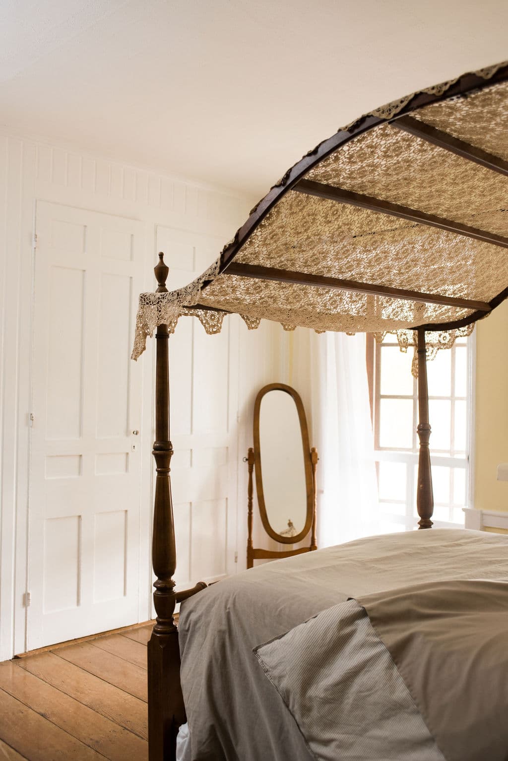 A wooden four-poster bed with a lace canopy in a softly lit bedroom featuring a mirror and white doors.