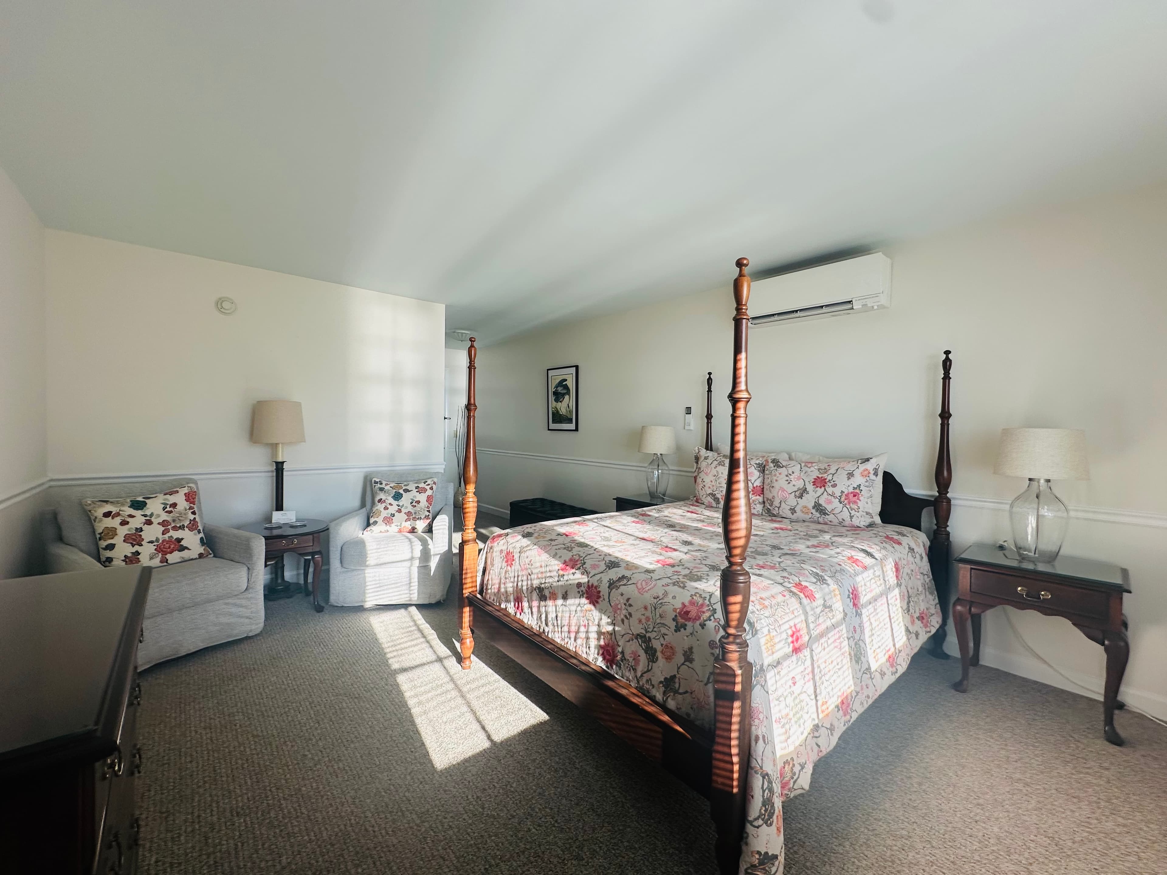 A cozy bedroom featuring a floral quilted bed, armchairs, and soft natural light.