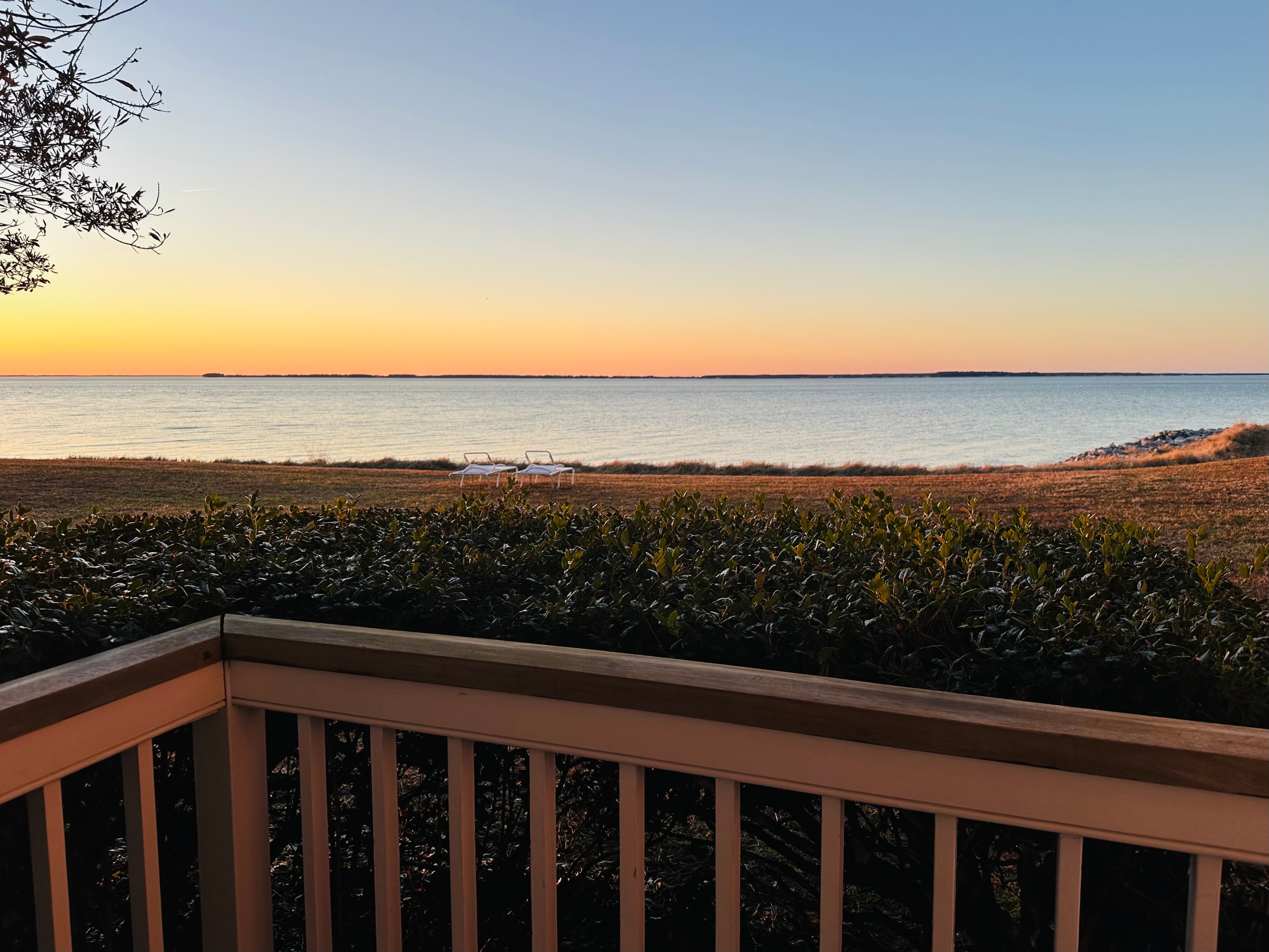Sunset over a calm lake, viewed from a balcony with bushes in the foreground.