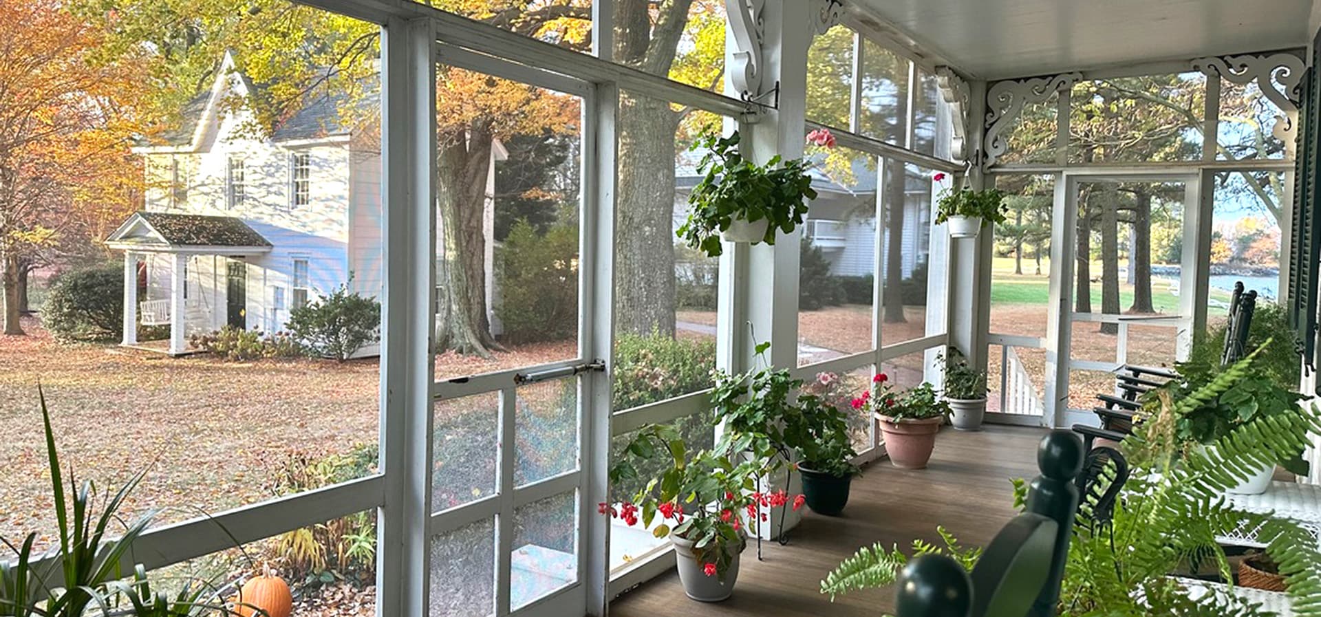 A cozy, screened porch filled with plants overlooks a colorful autumn landscape and a distant house.