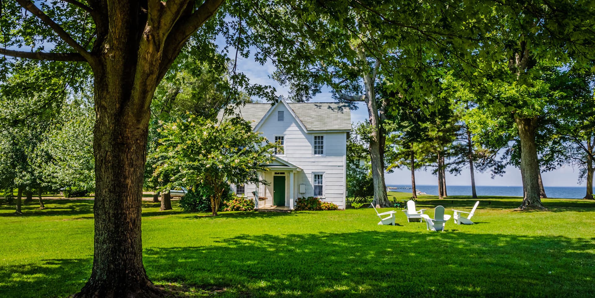 A white house surrounded by trees and a green lawn, with outdoor seating near a body of water.