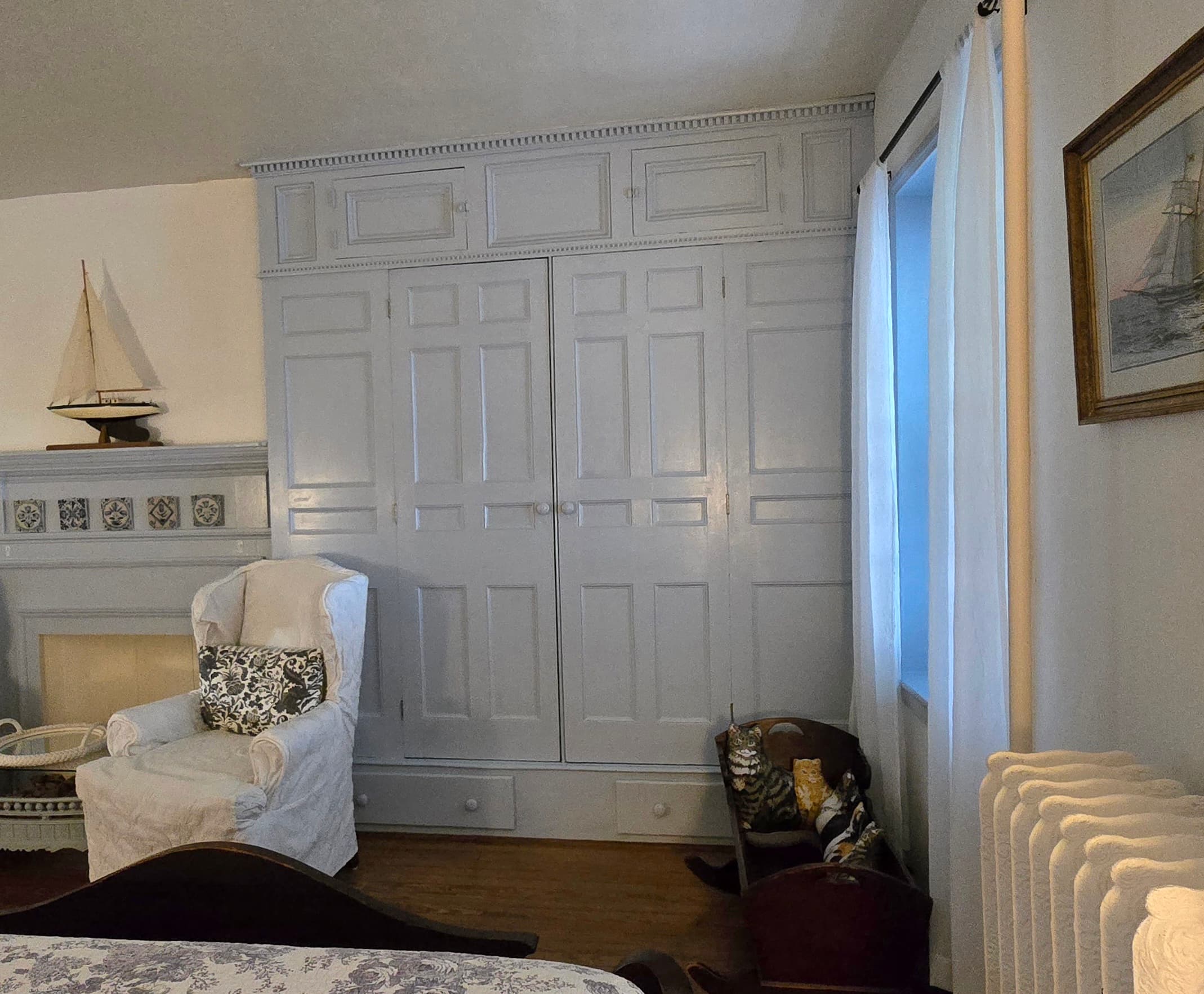 A cozy bedroom featuring a white armchair, built-in gray cabinets, and a window with sheer curtains.