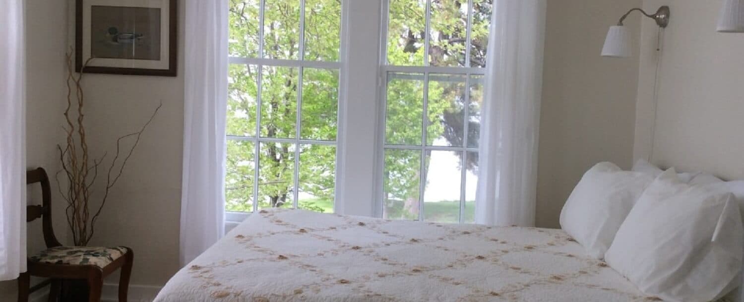 A bright, cozy bedroom featuring a bed with white linens and large windows overlooking greenery.