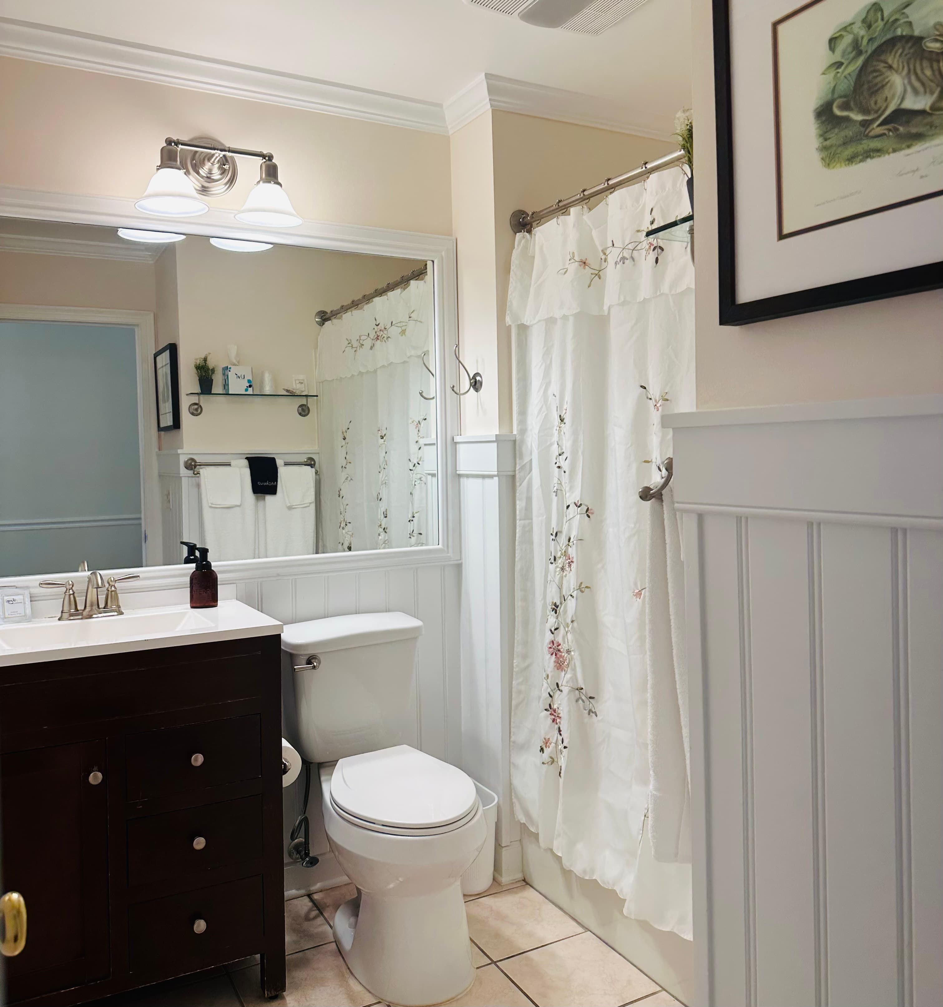 A compact bathroom featuring a wooden vanity, white toilet, floral shower curtain, and mirror with bright lighting.