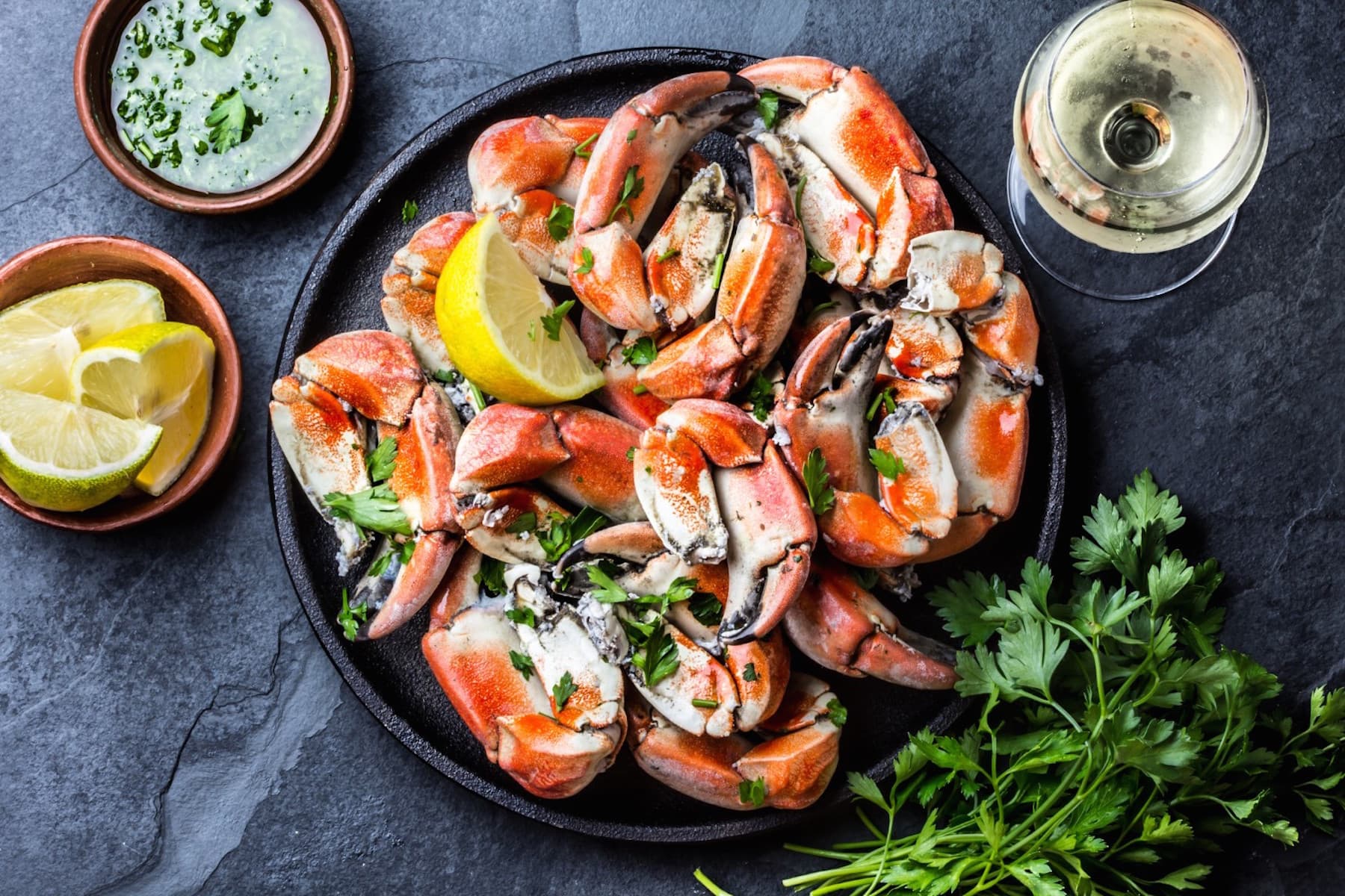 A platter of cooked crab claws garnished with lemon and parsley, accompanied by dipping sauce and a glass of white wine.