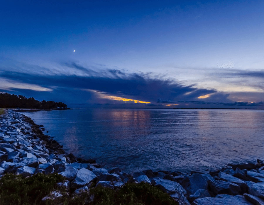 A tranquil shoreline at dusk with calm waters and a crescent moon above.