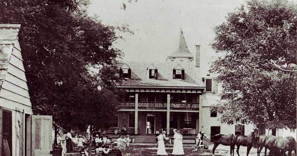 Historic photo of a group of people in front of a large building, with horses nearby.