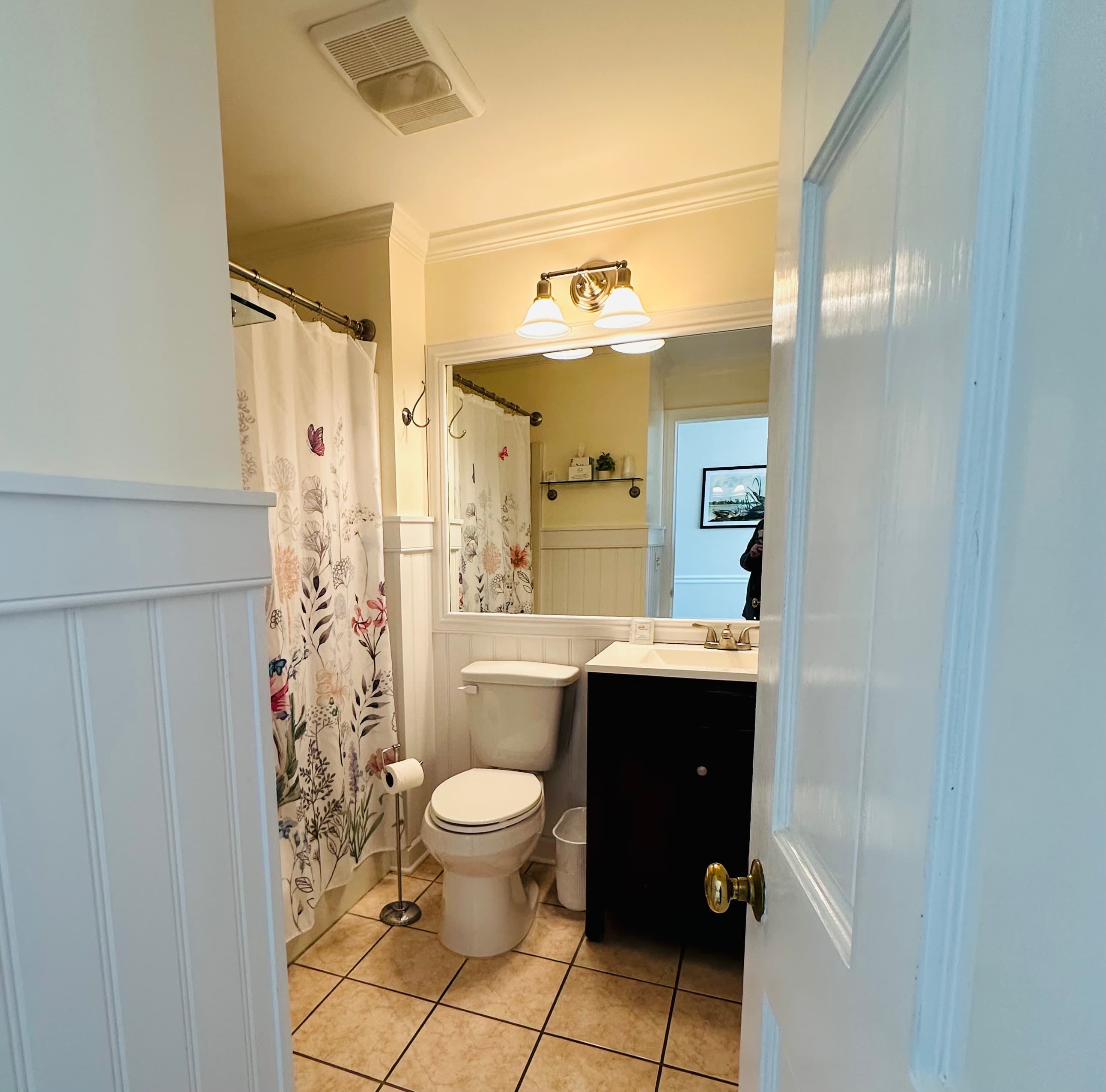 A bright bathroom featuring a floral shower curtain, sink, and toilet with tiled flooring.