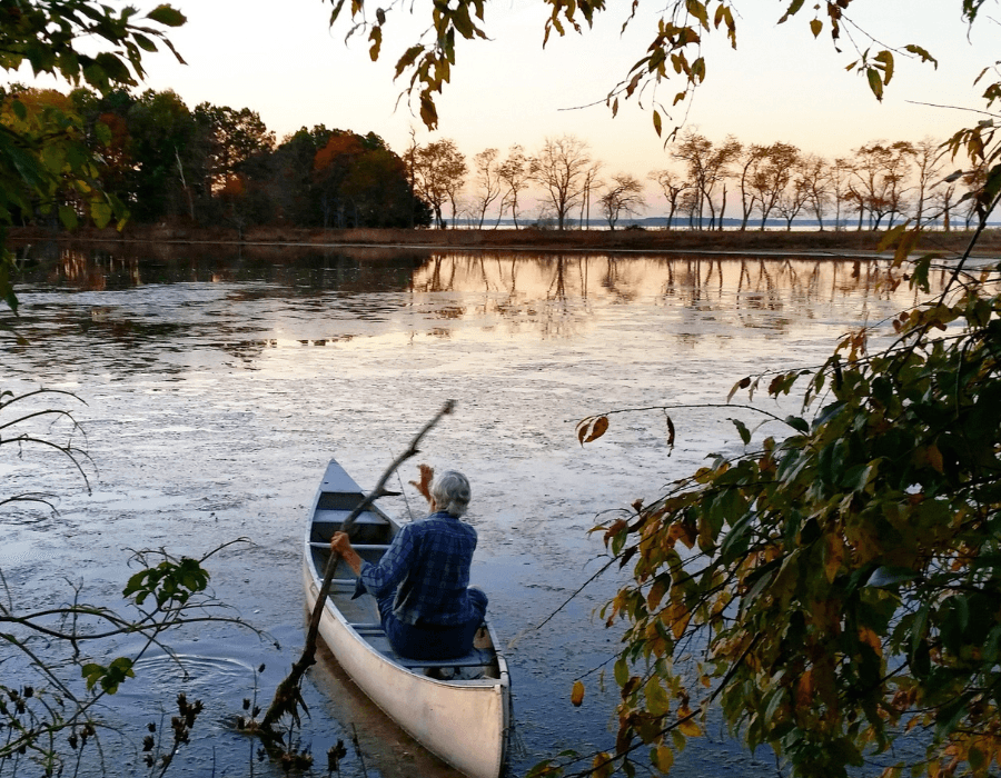 A person paddles a canoe on a tranquil lake at sunset, surrounded by trees.