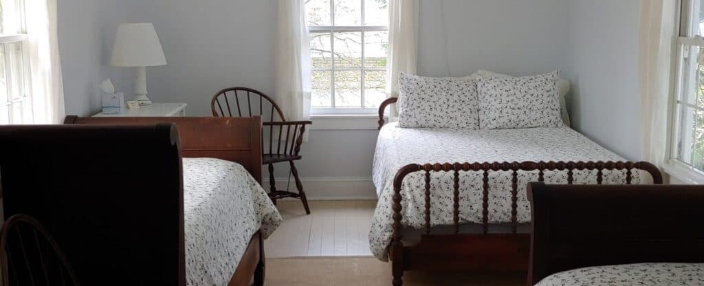 A cozy bedroom featuring two beds with floral bedding, a chair, and a lamp near a bright window.