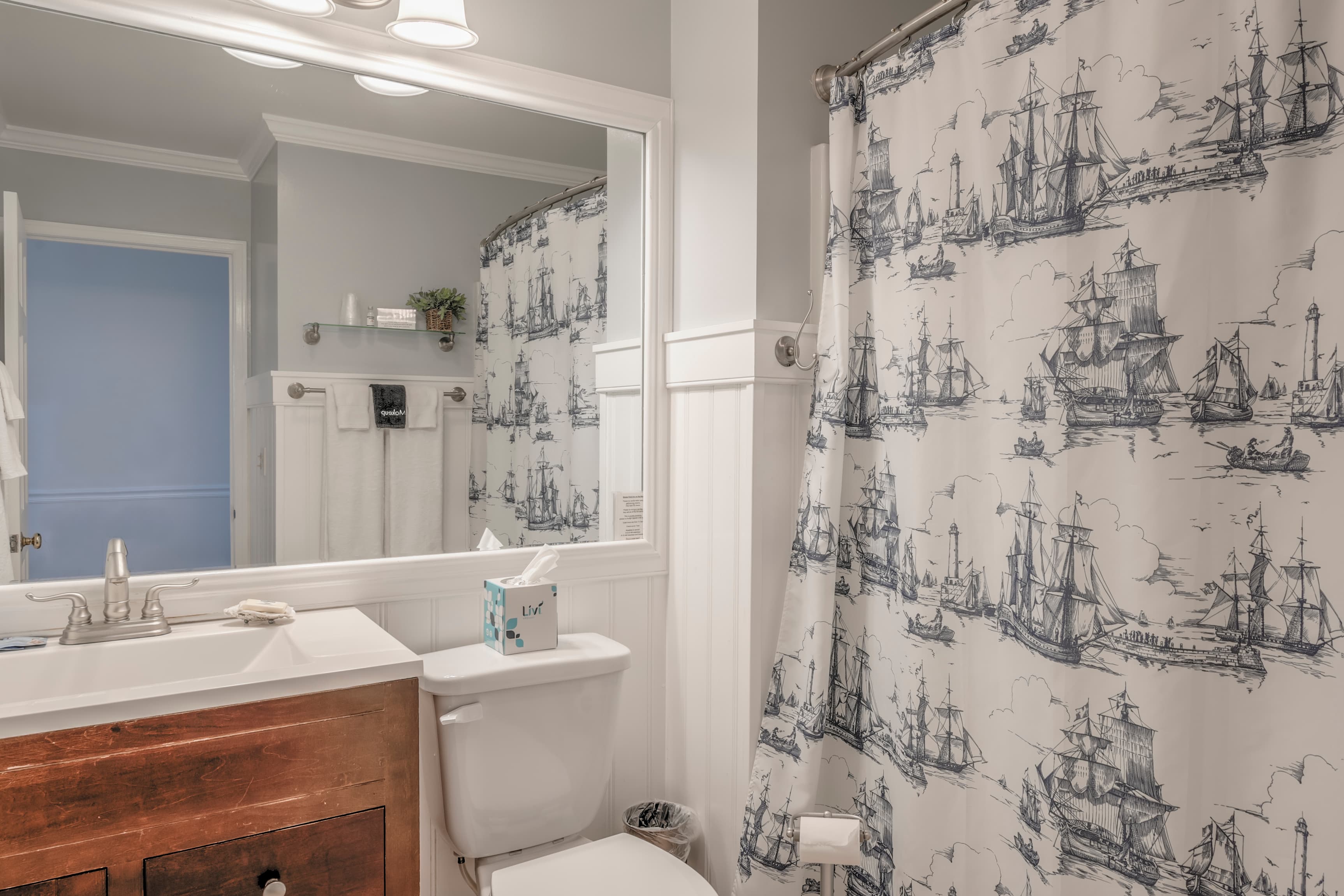 A bathroom featuring a ship-patterned shower curtain, sink with wooden vanity, and a large mirror.