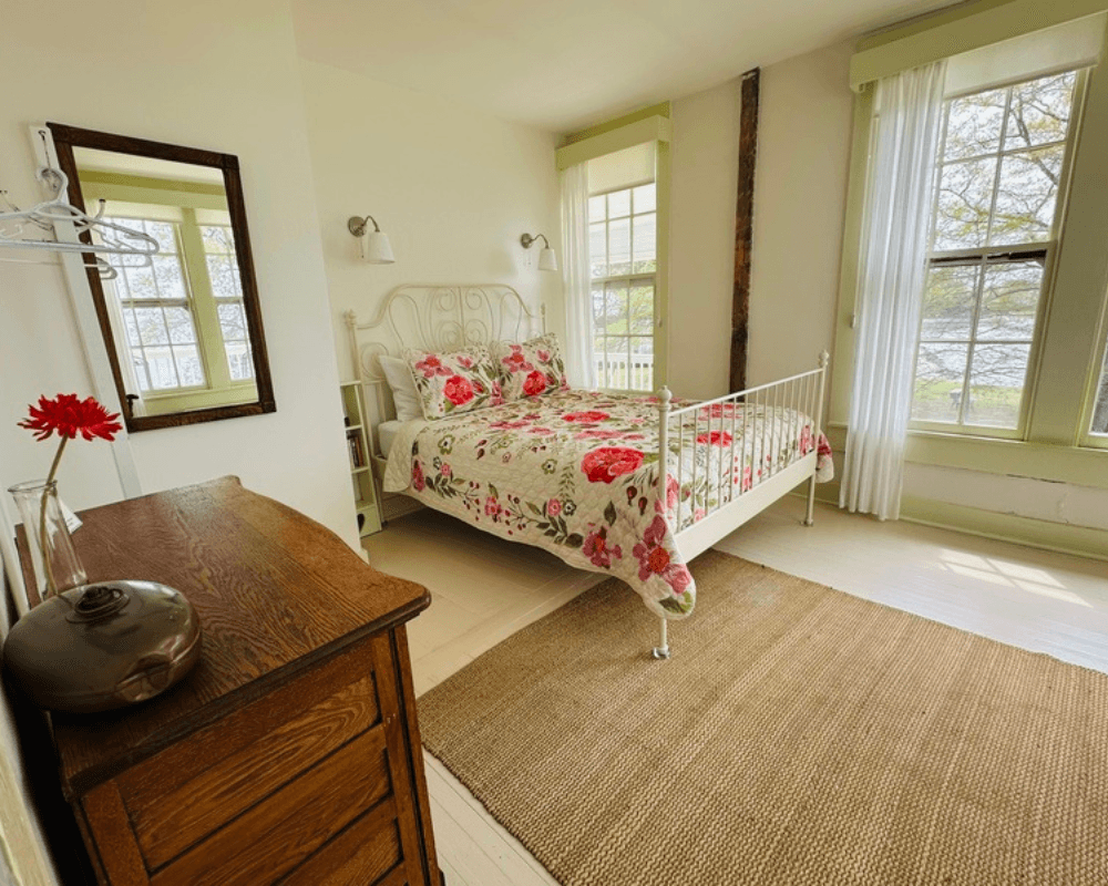 A bright bedroom featuring a white metal bed with floral bedding, a wooden dresser, and large windows overlooking greenery.