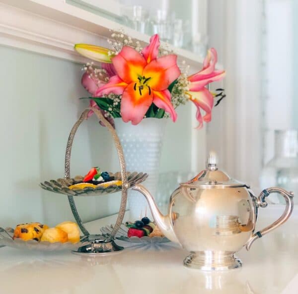 A silver teapot sits beside a tiered stand of fruit and a vase of pink lilies.
