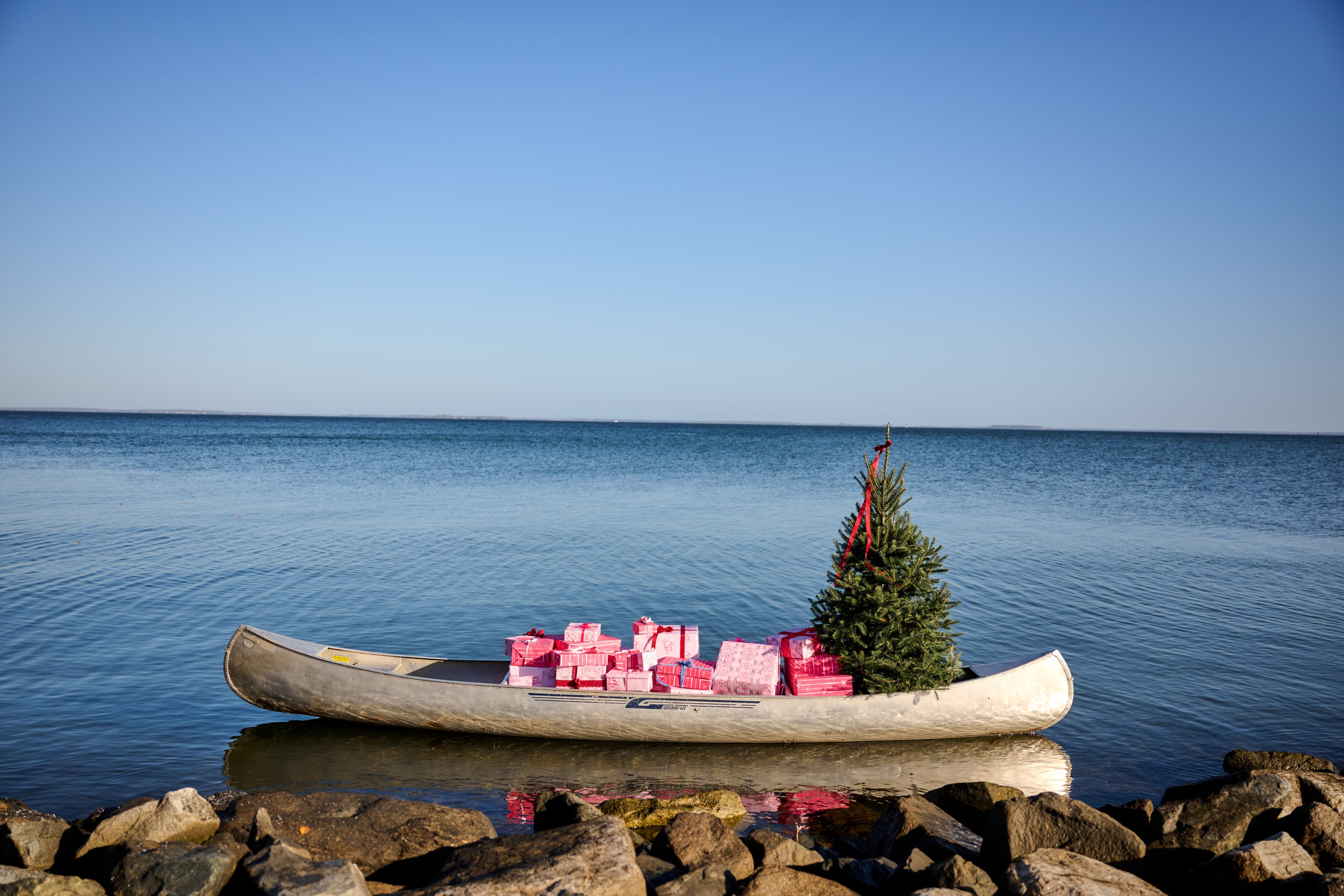 A canoe decorated with a Christmas tree and pink gift boxes floats on calm water against a clear blue sky.