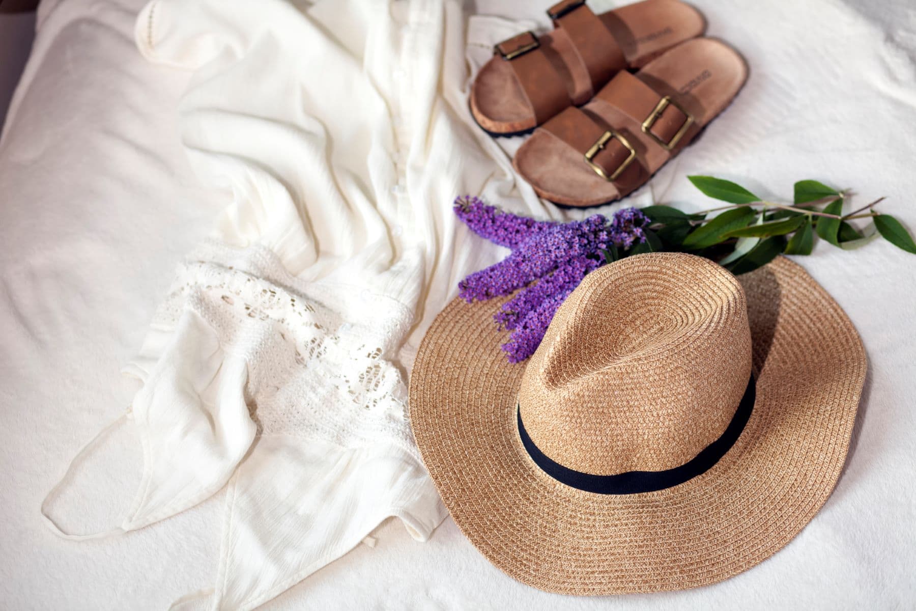 A flat lay featuring a white dress, brown sandals, a straw hat, purple flowers, and green leaves. A flat lay featuring a white dress, brown sandals, a straw hat, purple flowers, and green leaves.