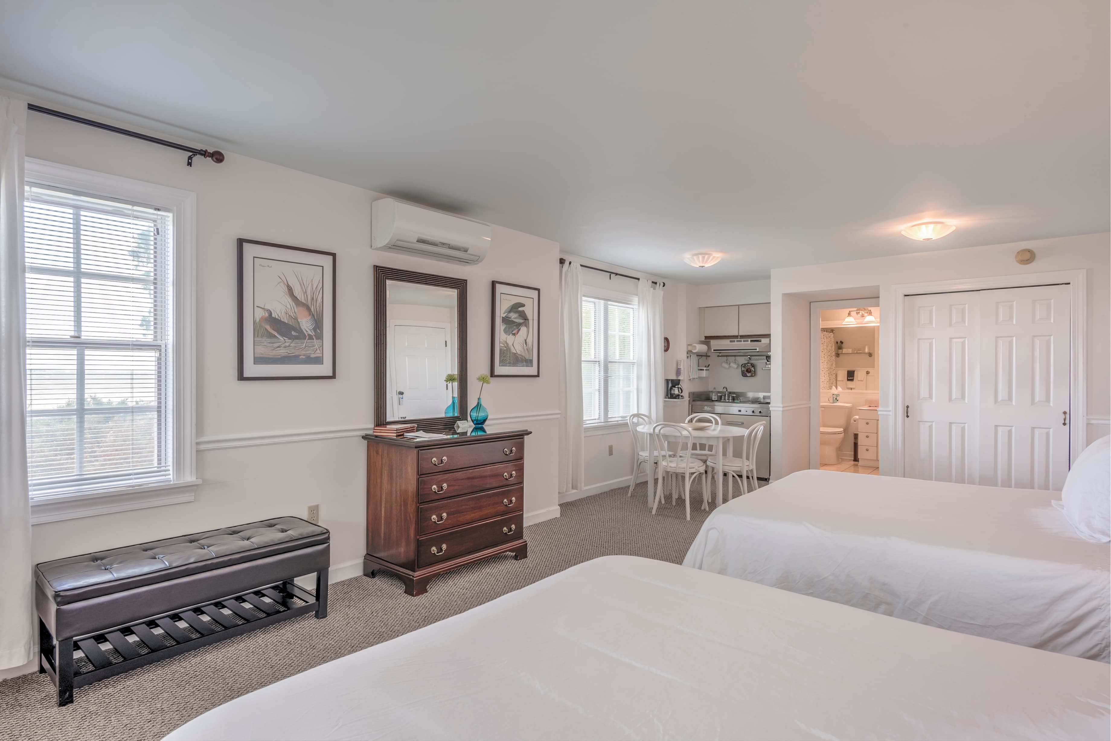 A bright, airy hotel room featuring two beds, a wooden dresser, and a kitchenette.