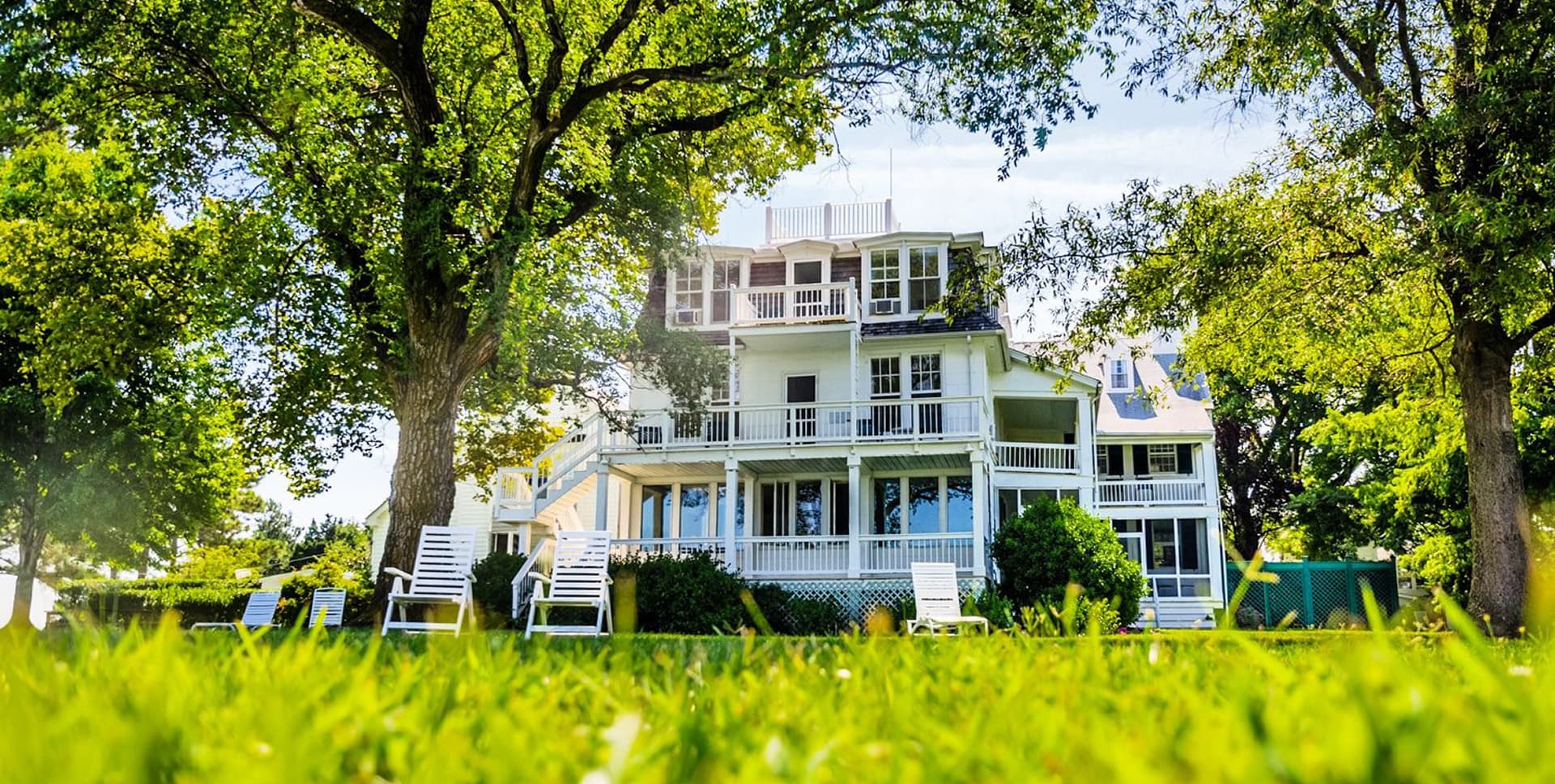 A large, charming house surrounded by trees and green grass, featuring a porch and white lounge chairs.