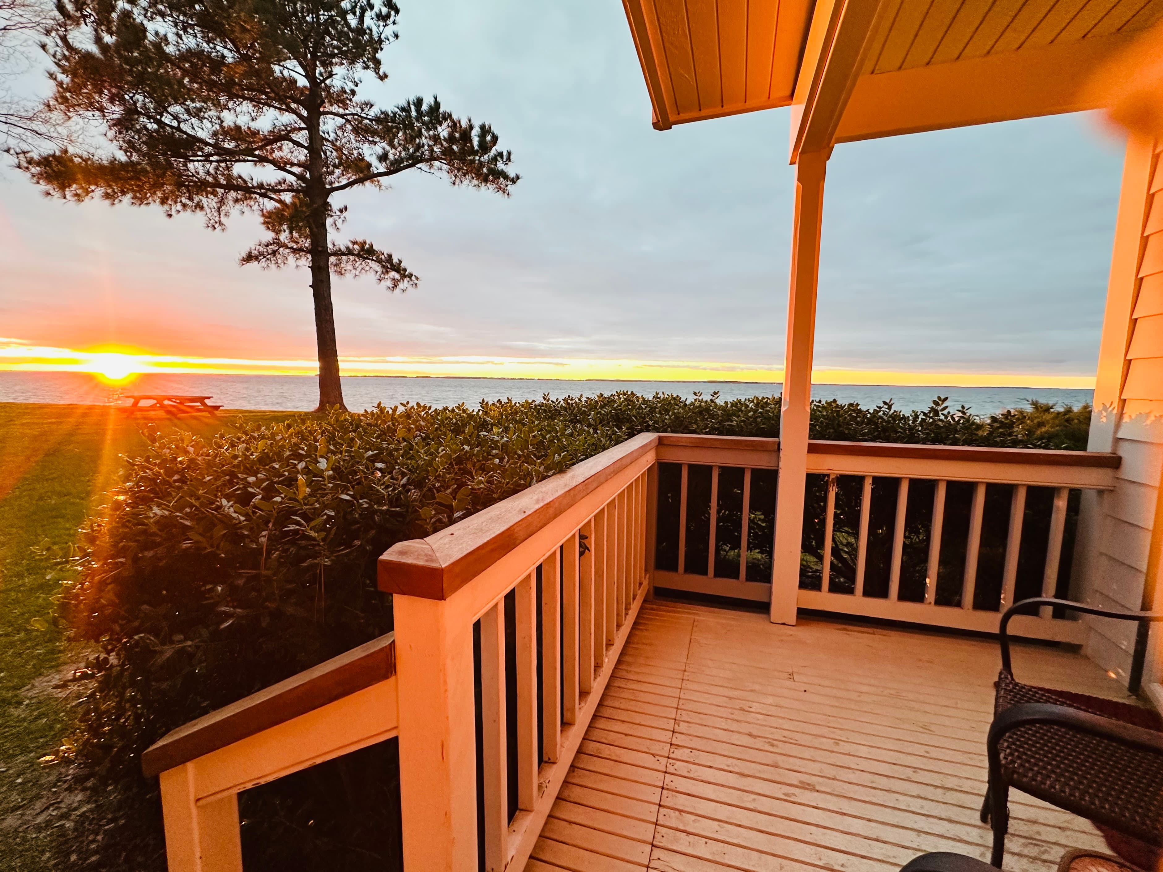 A wooden porch overlooking a sunset over a body of water, framed by trees and greenery.