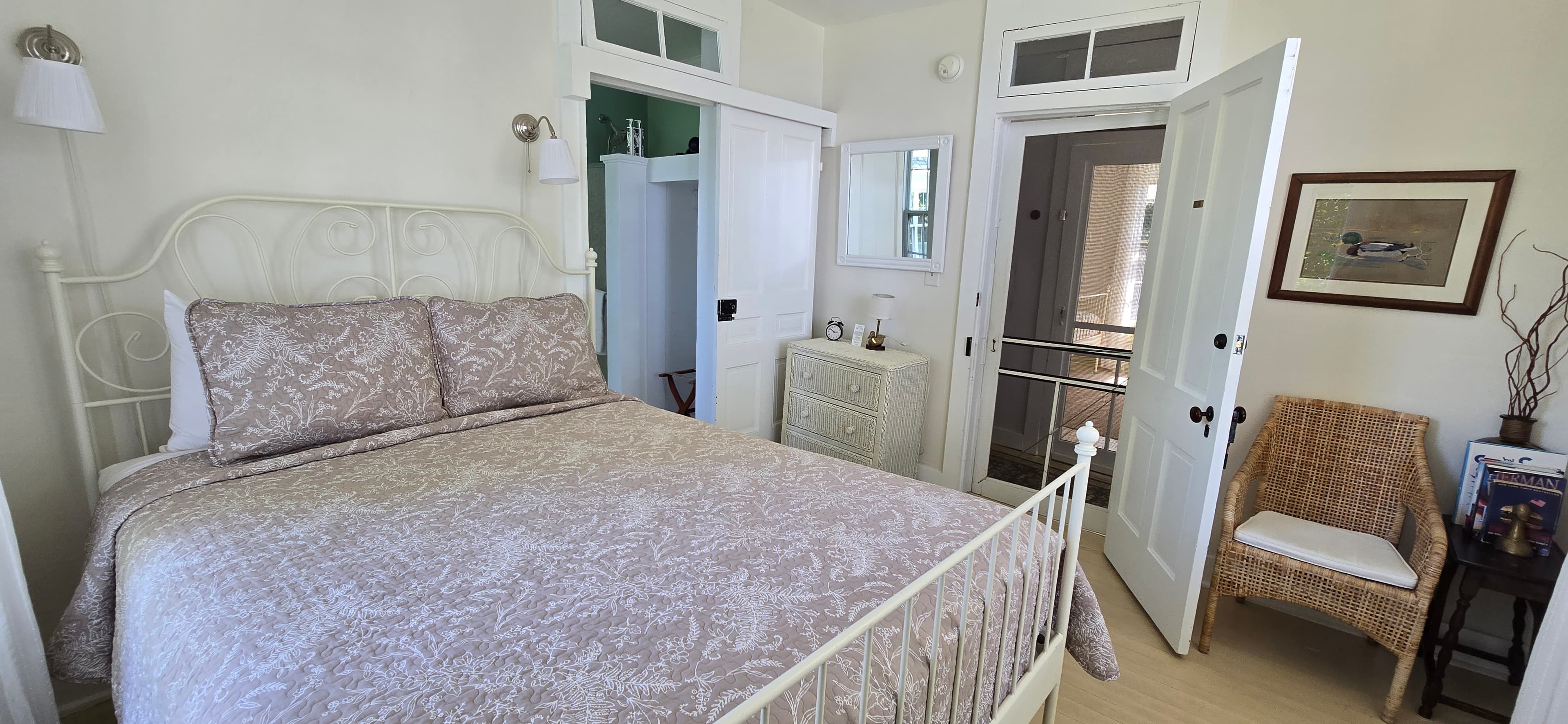 A bright bedroom featuring a white bed with patterned bedding, a wicker chair, and open doors to a hallway.