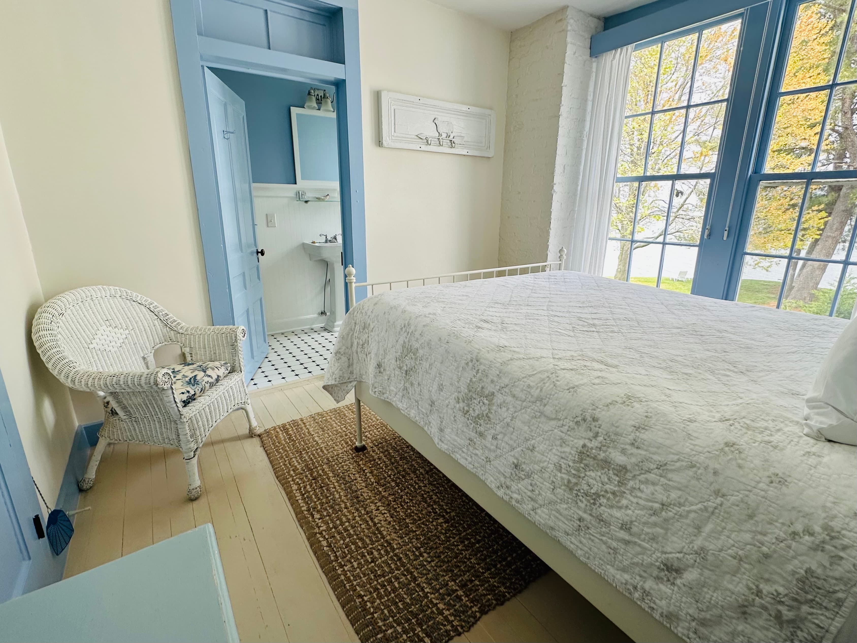 A bright, airy bedroom featuring a white bedspread, wicker chair, and large windows with blue trim.