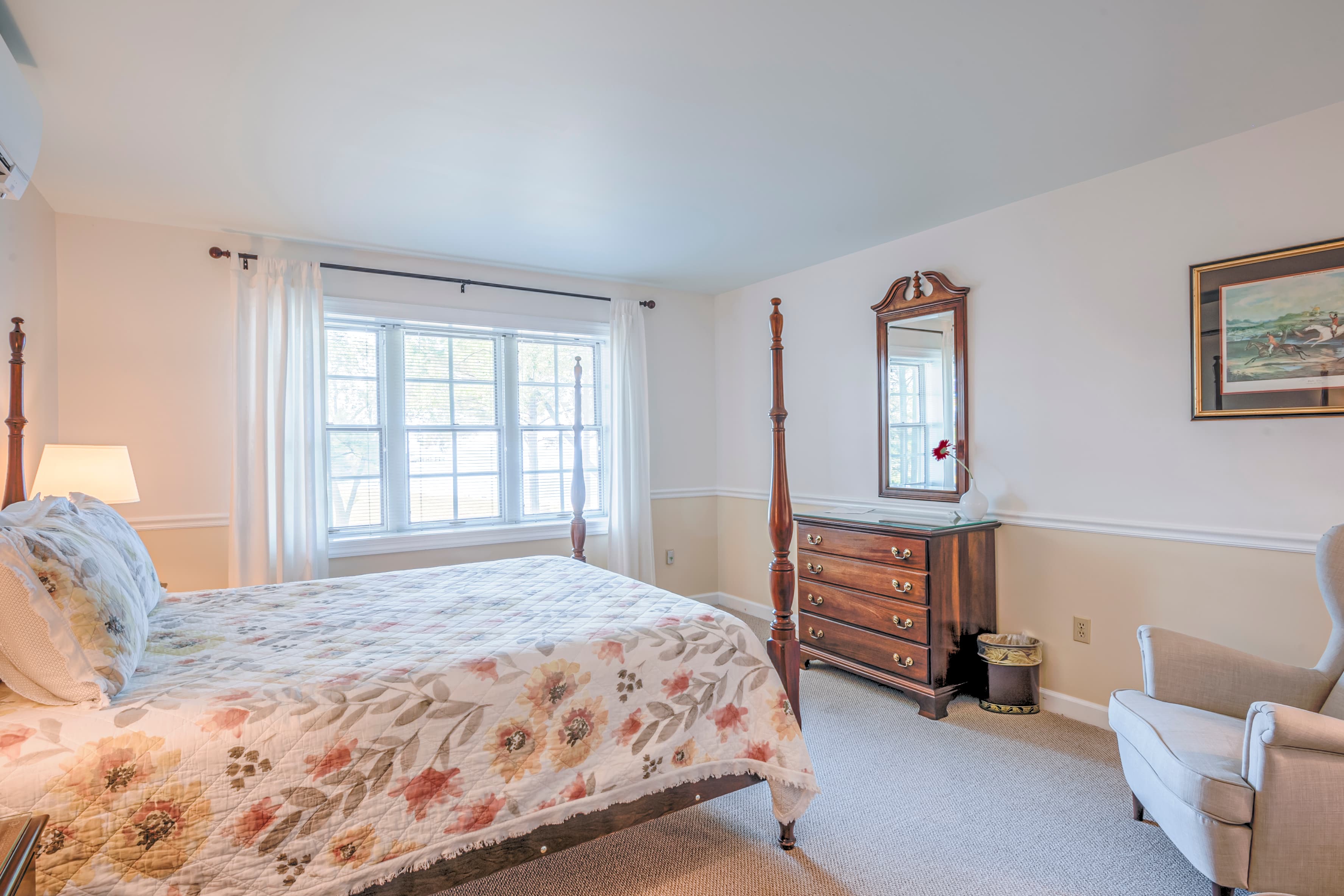 A cozy bedroom featuring a floral quilted bedspread, a wooden dresser with a mirror, and a plush armchair beside a large window.