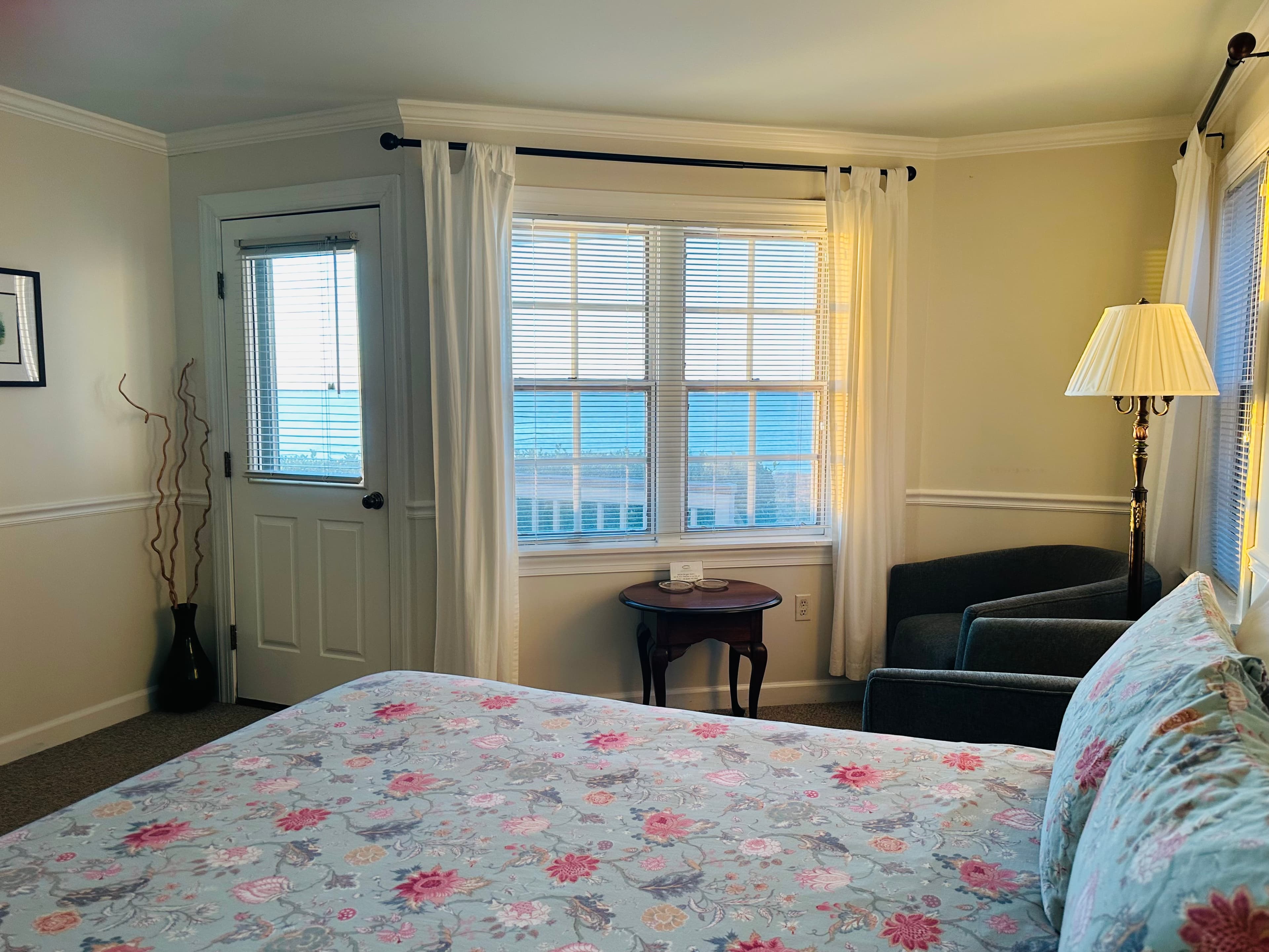 Bright bedroom with a floral bedspread, a comfortable chair, and a window offering a view of the ocean.