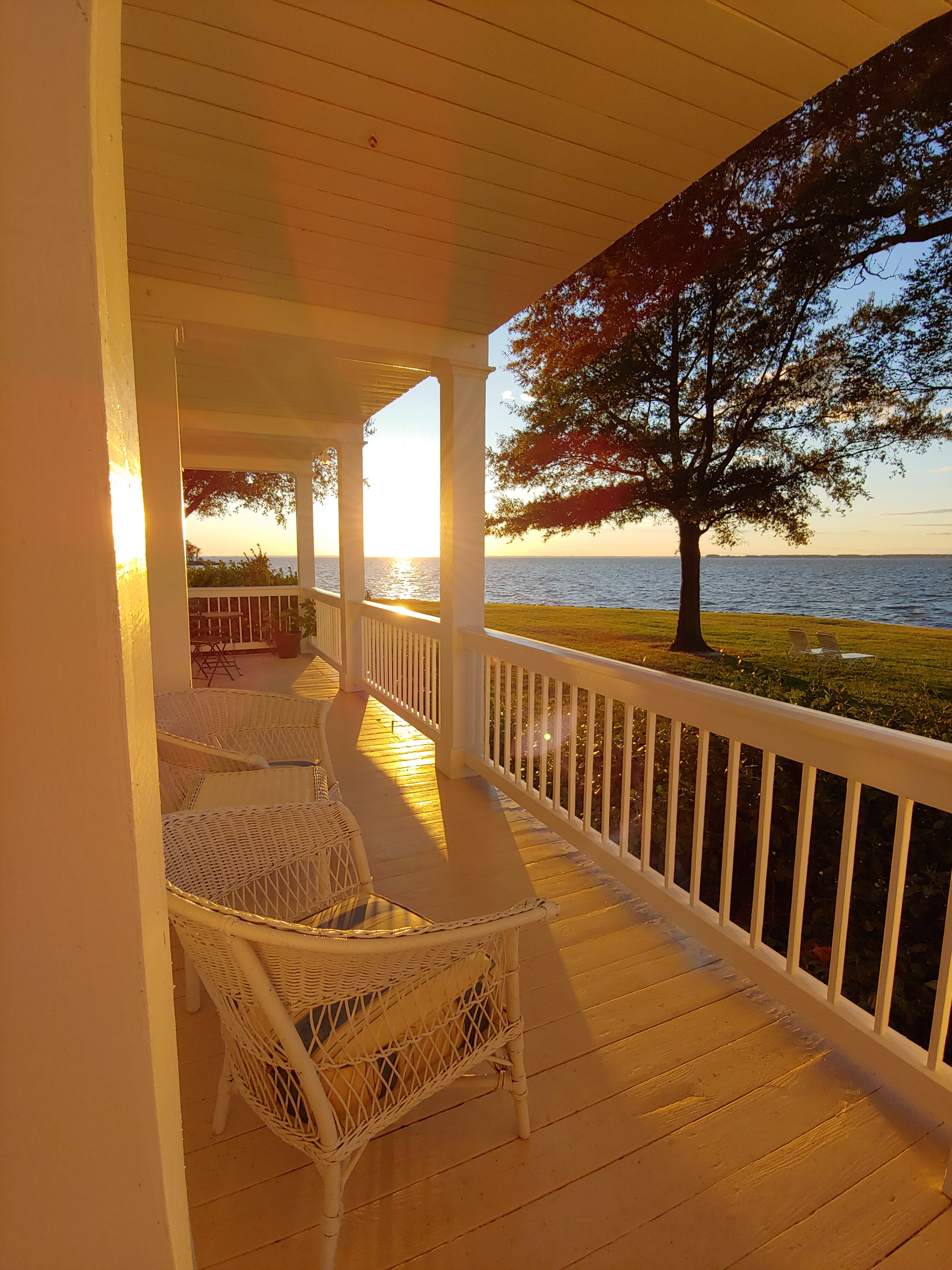 A sunlit porch with wicker chairs overlooks a tranquil lake at sunset.