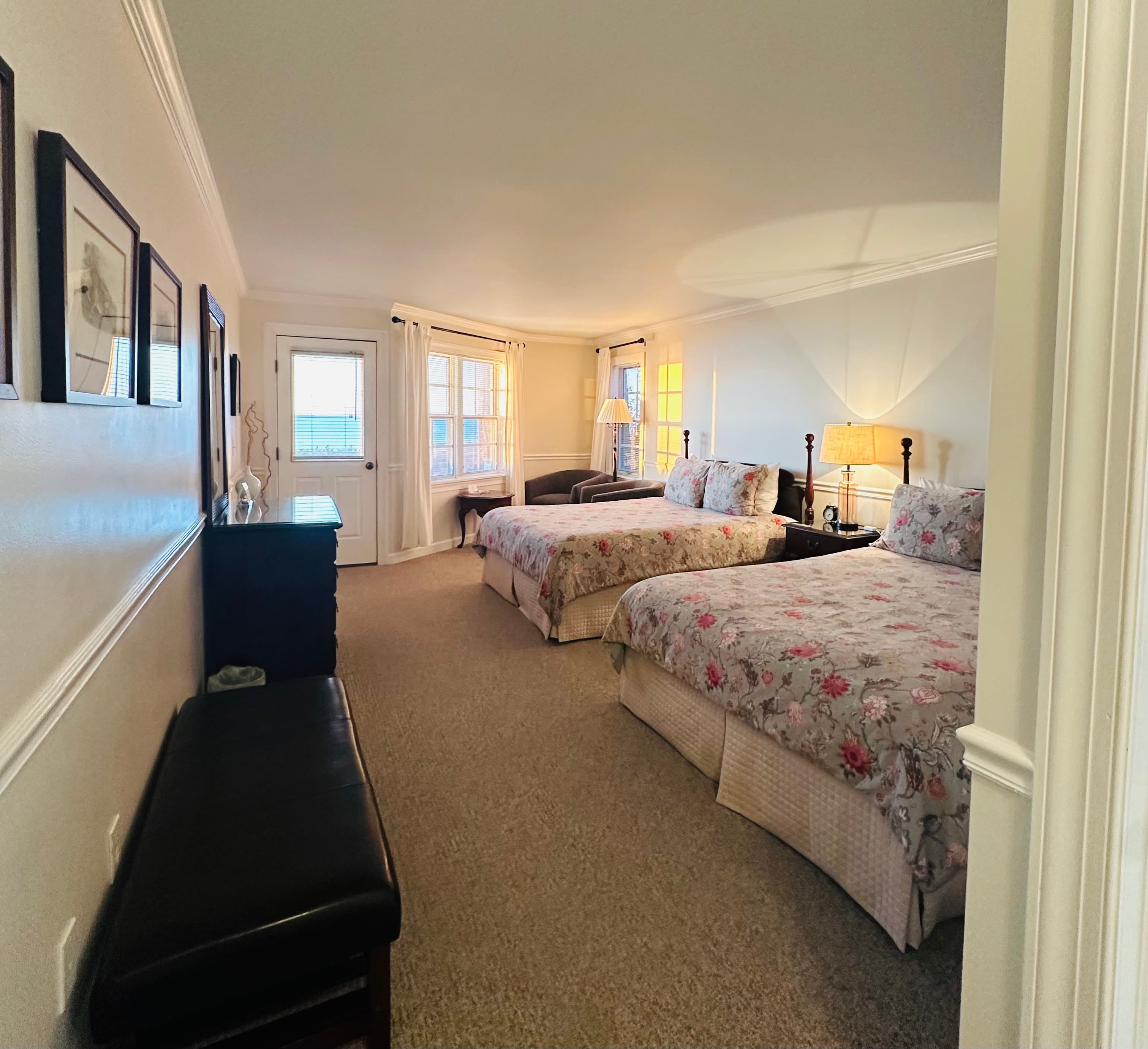 A cozy guest room featuring two floral-patterned beds, a small seating area, and large windows with natural light.