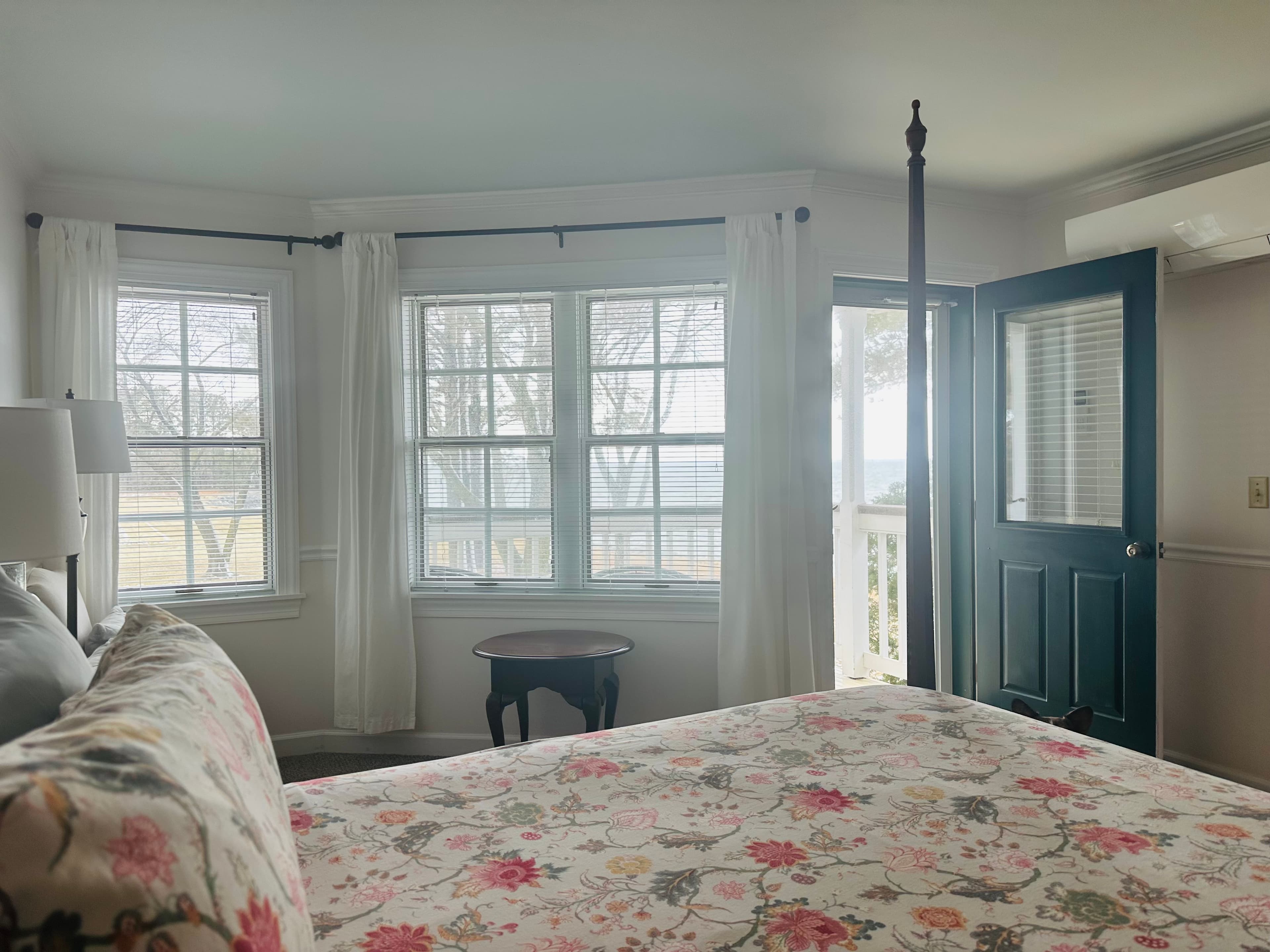 A cozy bedroom with a floral-patterned bedspread, featuring large windows and a door leading to an outside view.