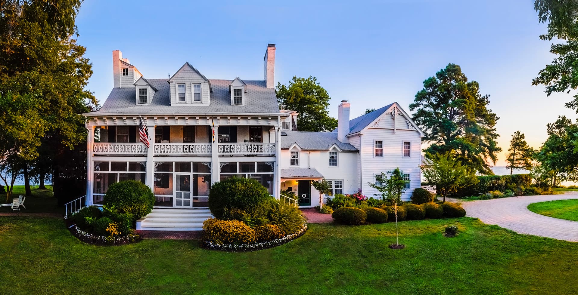 A large white house with a wraparound porch surrounded by well-maintained gardens and trees.