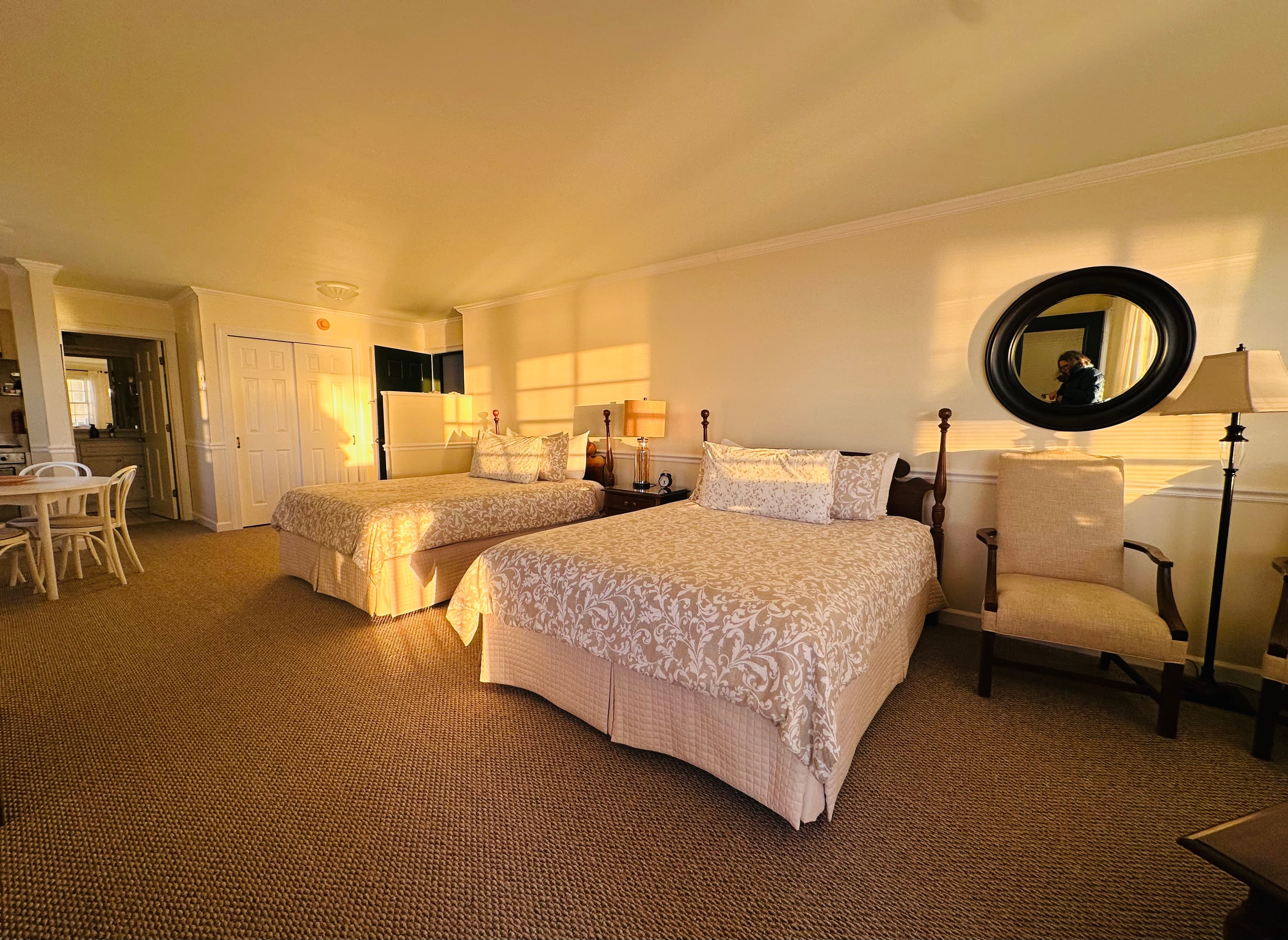 A sunlit hotel room featuring two queen beds, a mirror, and a small dining area.