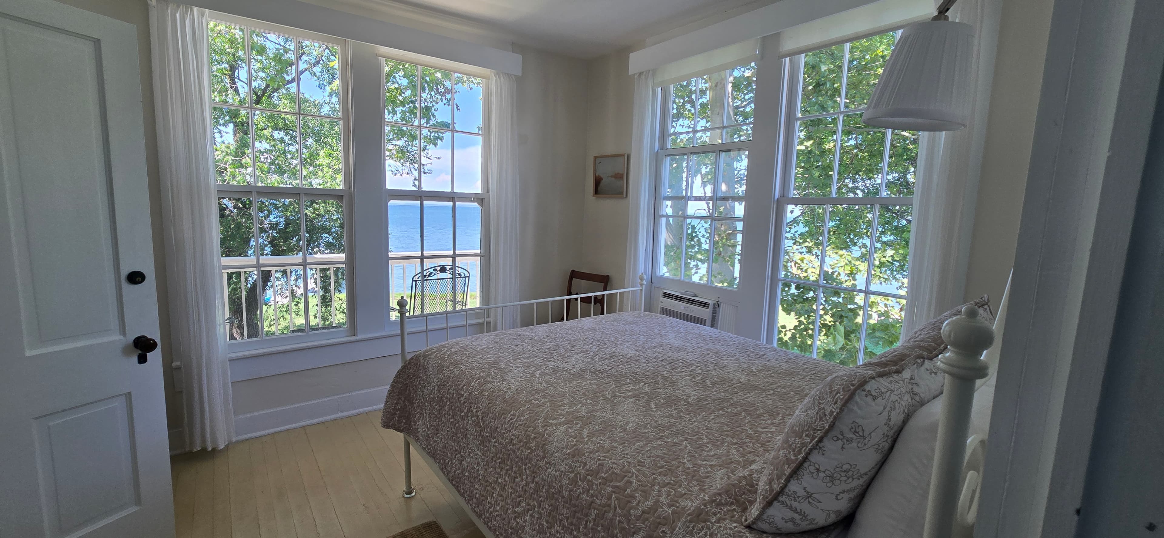 Bright bedroom with a white bed and large windows overlooking a tree-lined view and water.