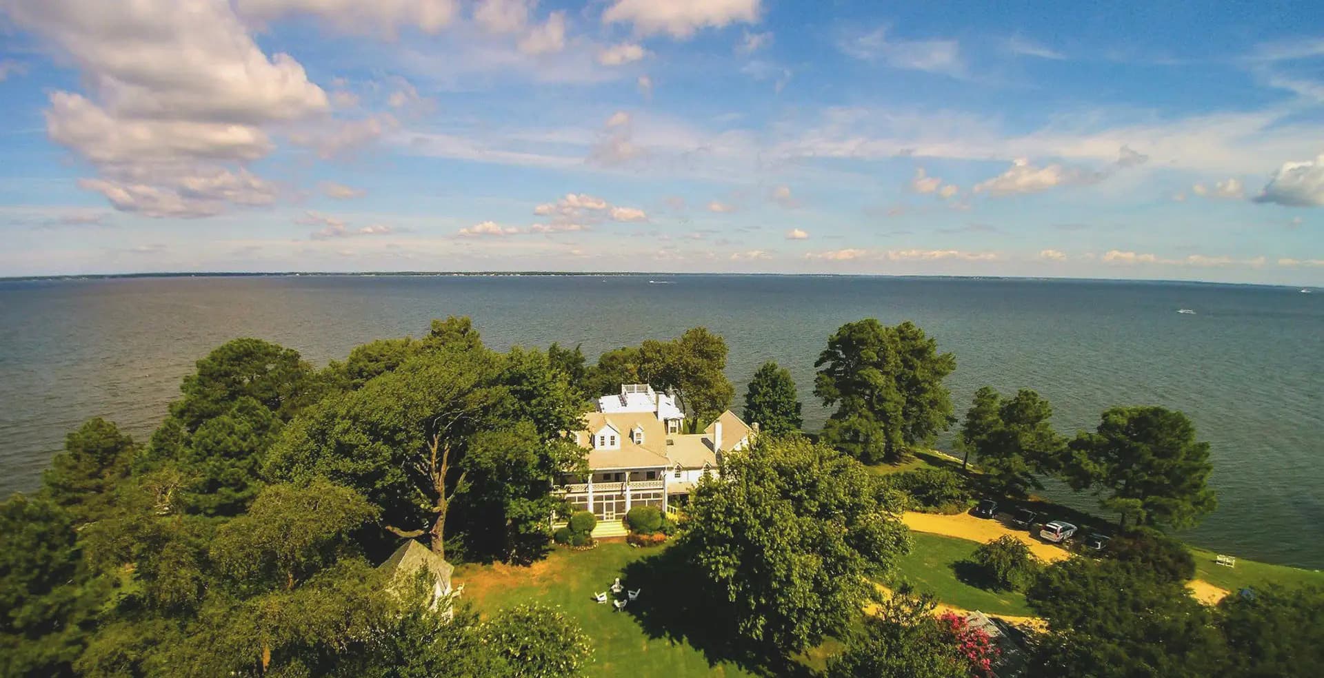 Aerial view of a waterfront house surrounded by trees and a calm body of water under a blue sky with clouds.