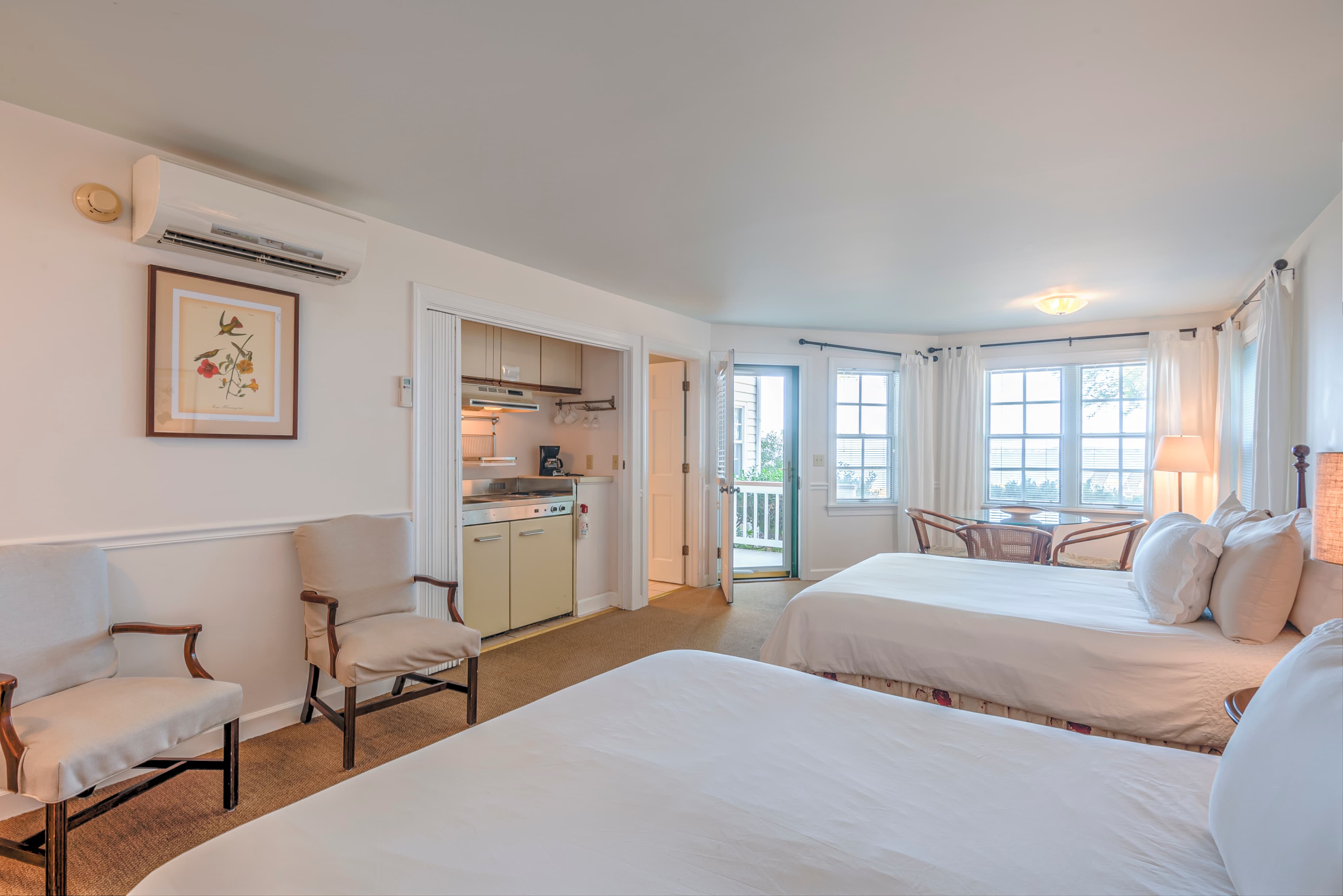 A bright hotel room featuring two beds, a small kitchenette, and a sliding glass door leading to a balcony with a view.