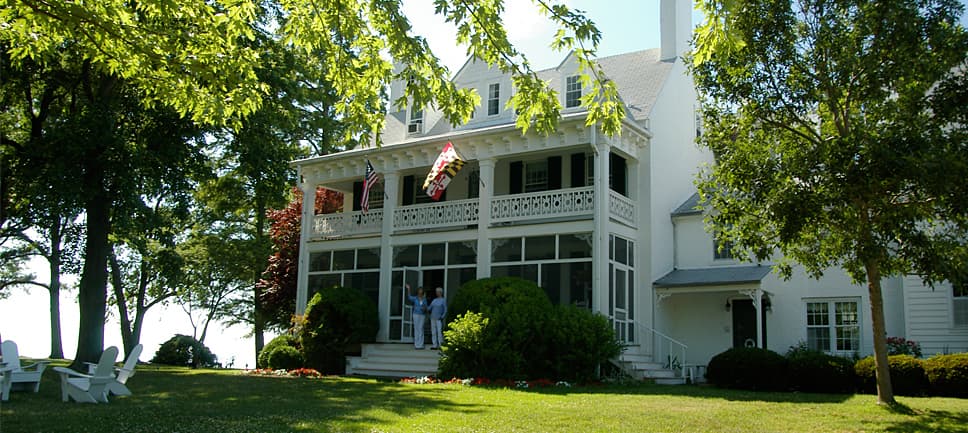 White house with a spacious porch surrounded by greenery and flags.