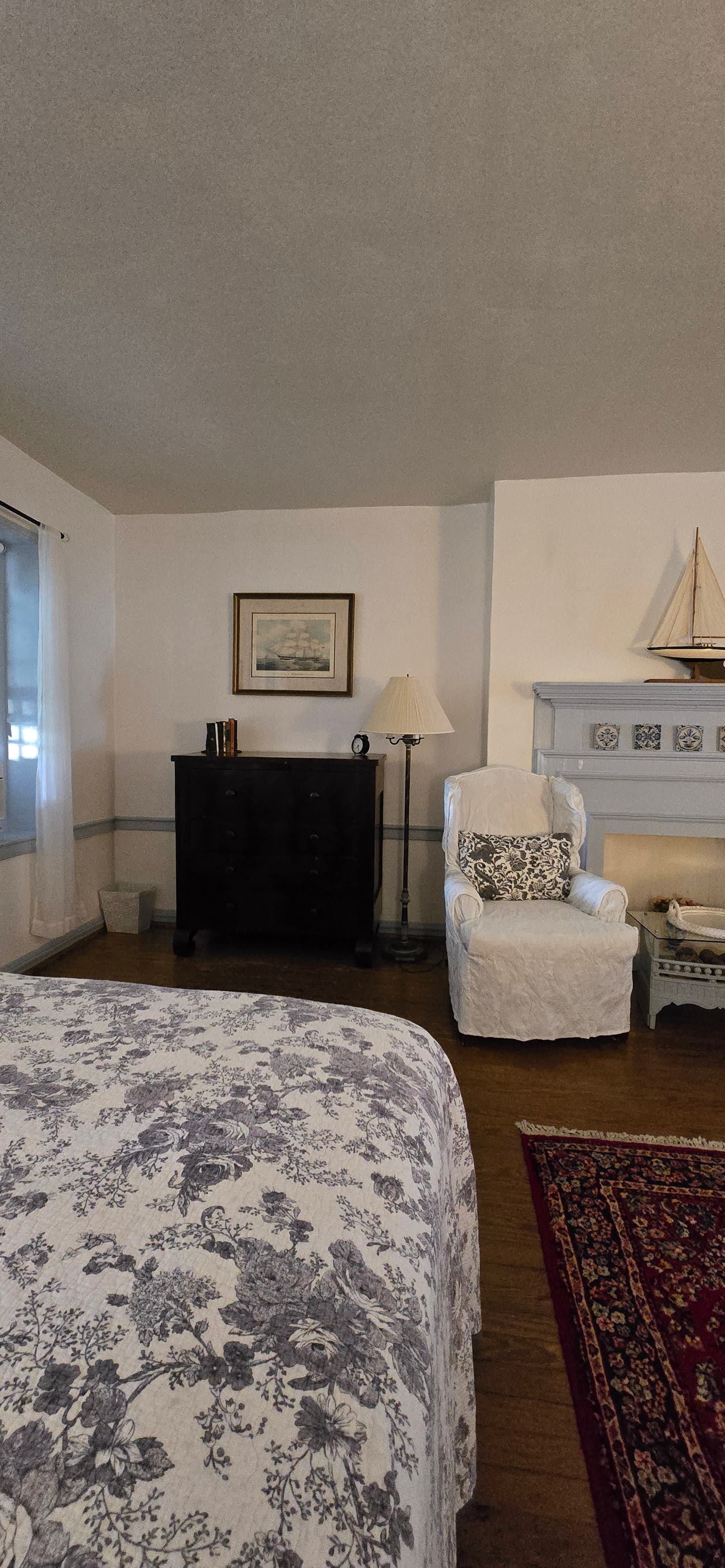 A cozy bedroom featuring a floral bedspread, an armchair, and a lamp beside a dark wooden dresser.
