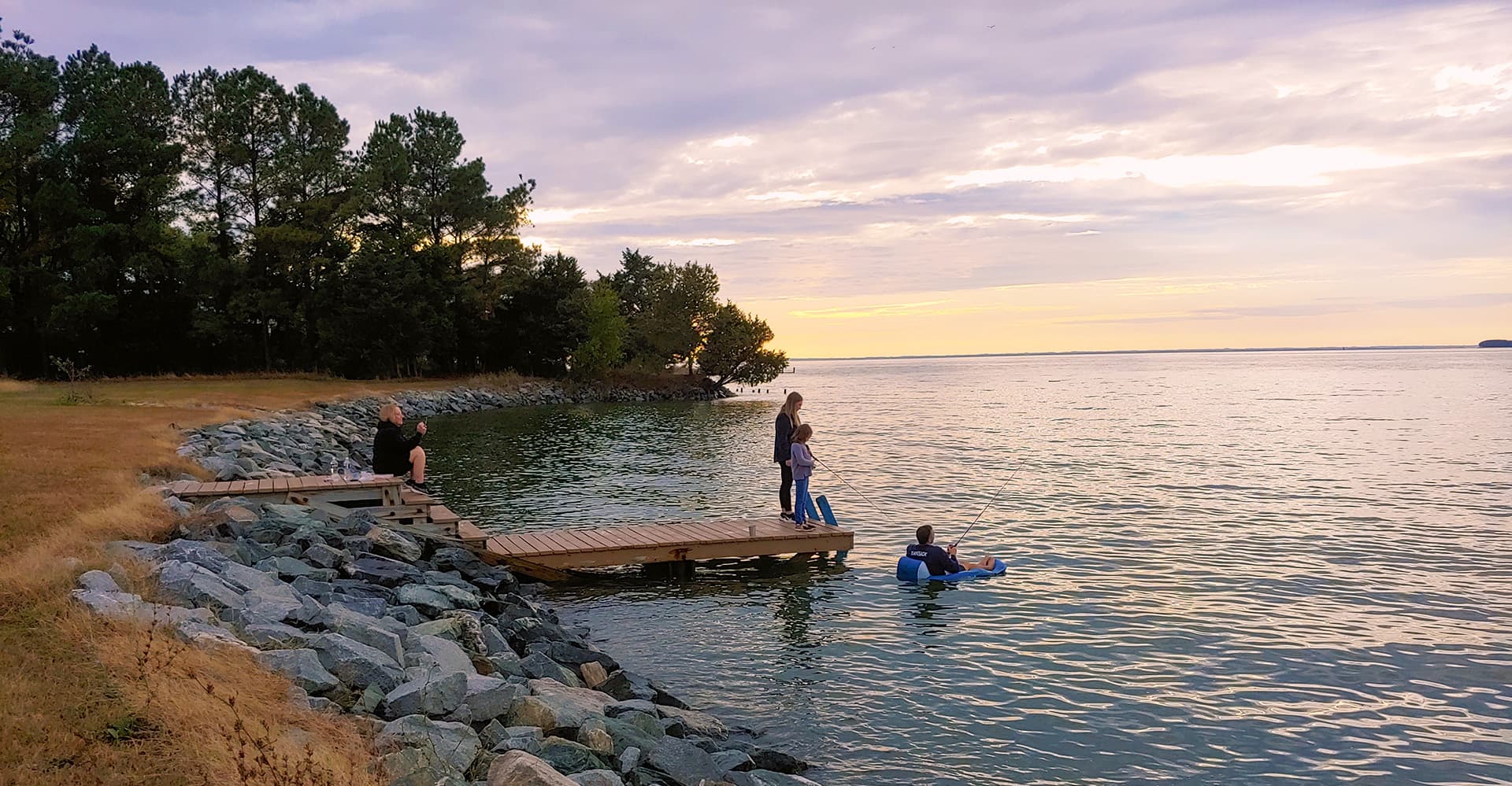 People enjoy a tranquil lakeside scene at sunset, with one person in the water and others on the dock.