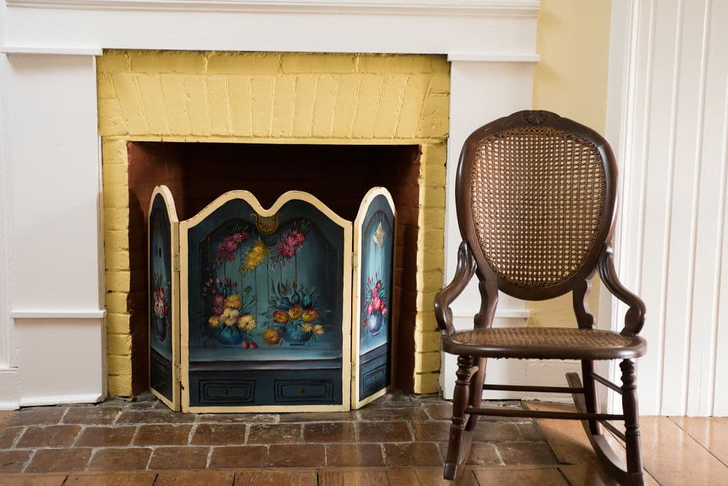 A vintage decorative fireplace screen stands next to a wooden caned chair in a cozy room.