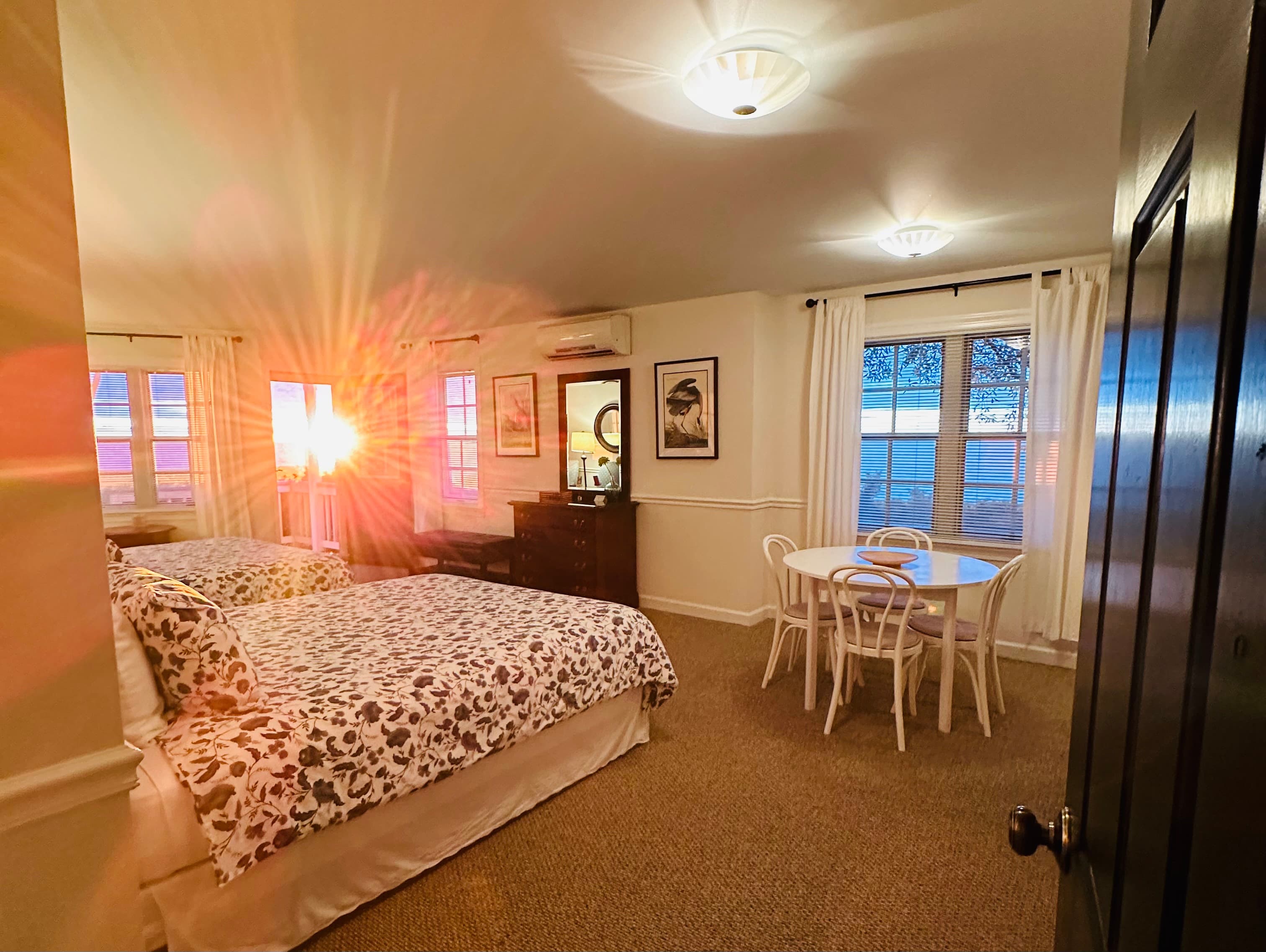 A cozy hotel room with two beds, a small dining table, and sunlight streaming through the windows.