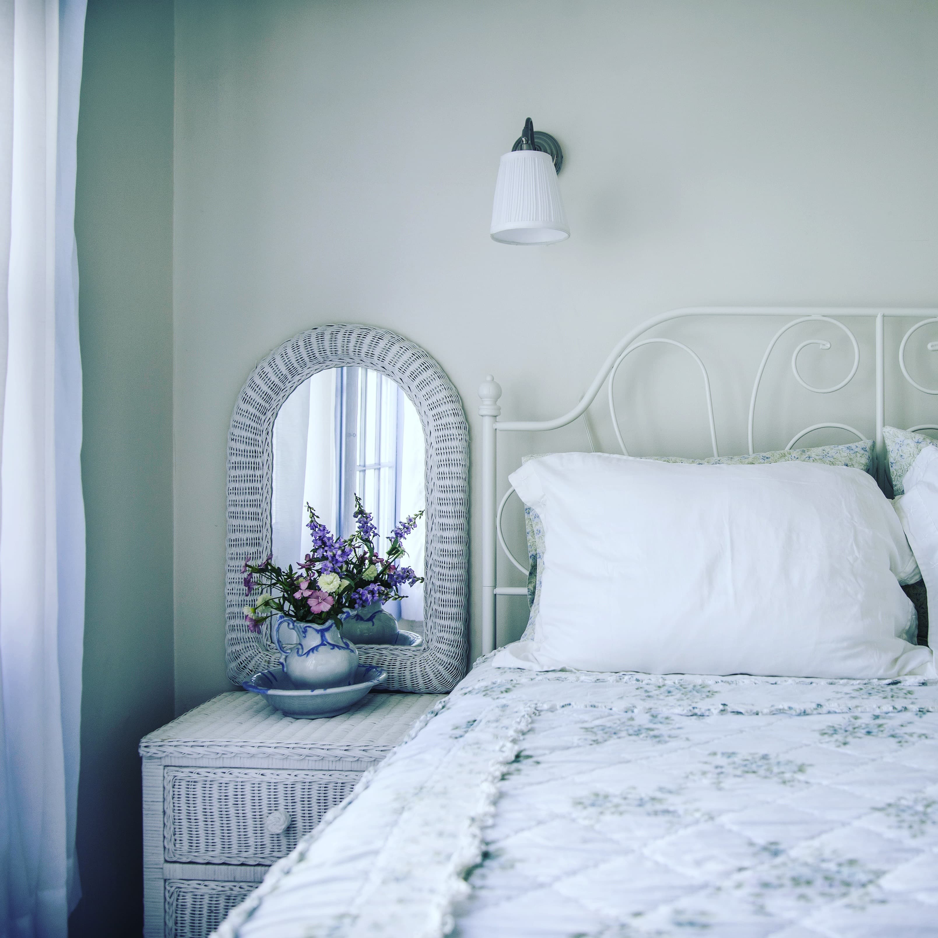 A cozy bedroom featuring a white metal bed, floral bedding, and a bedside table with a mirror and a vase of flowers.