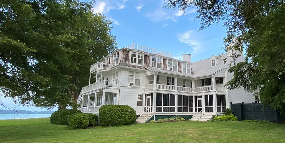 A large white house with multiple balconies surrounded by green lawns and trees, overlooking a body of water.