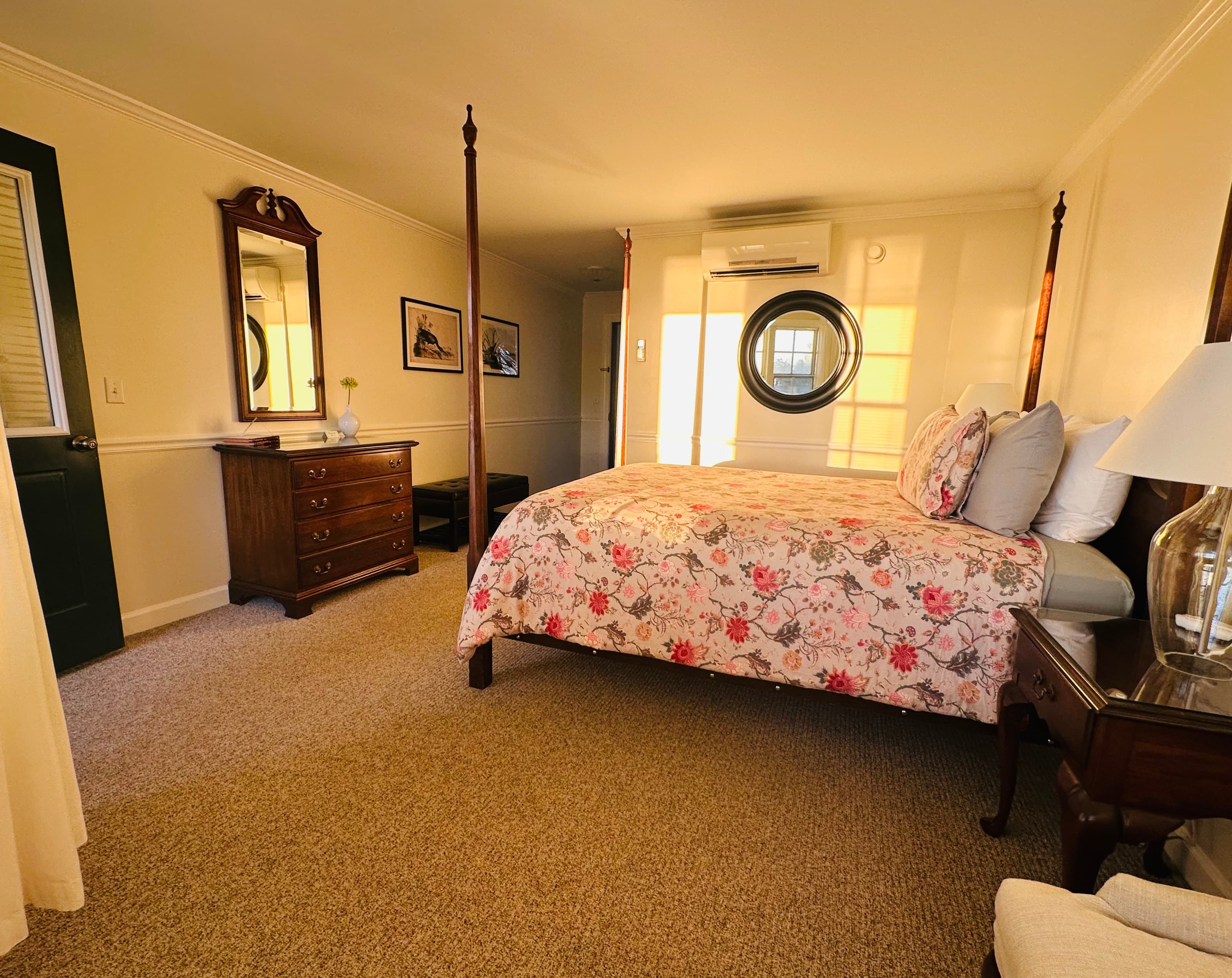 A cozy bedroom featuring a floral bedspread, a mirror, a dresser, and a round window.