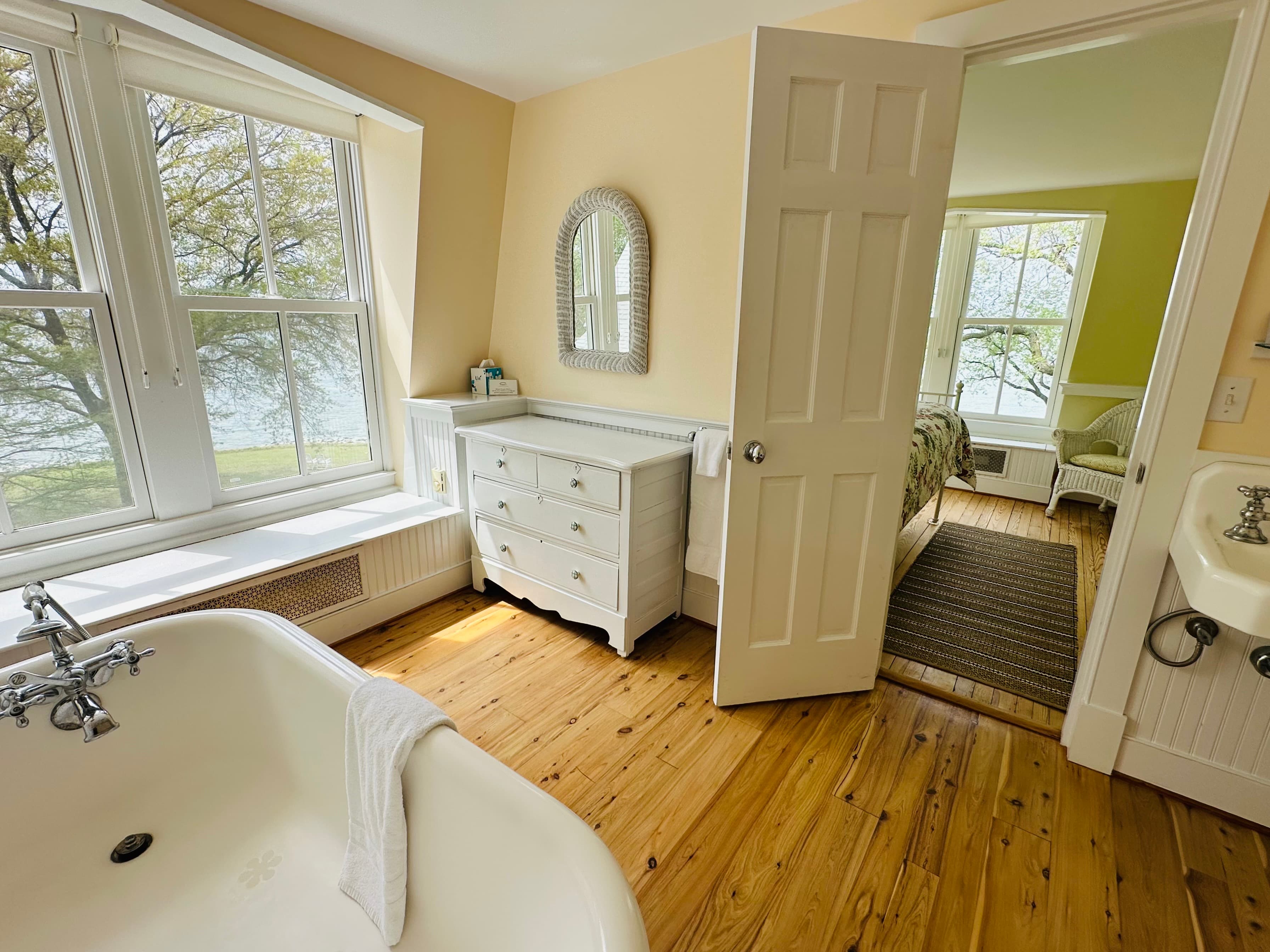 A bright bathroom featuring a vintage bathtub, a white dresser, and a doorway leading to a cozy bedroom with views of the outdoors.